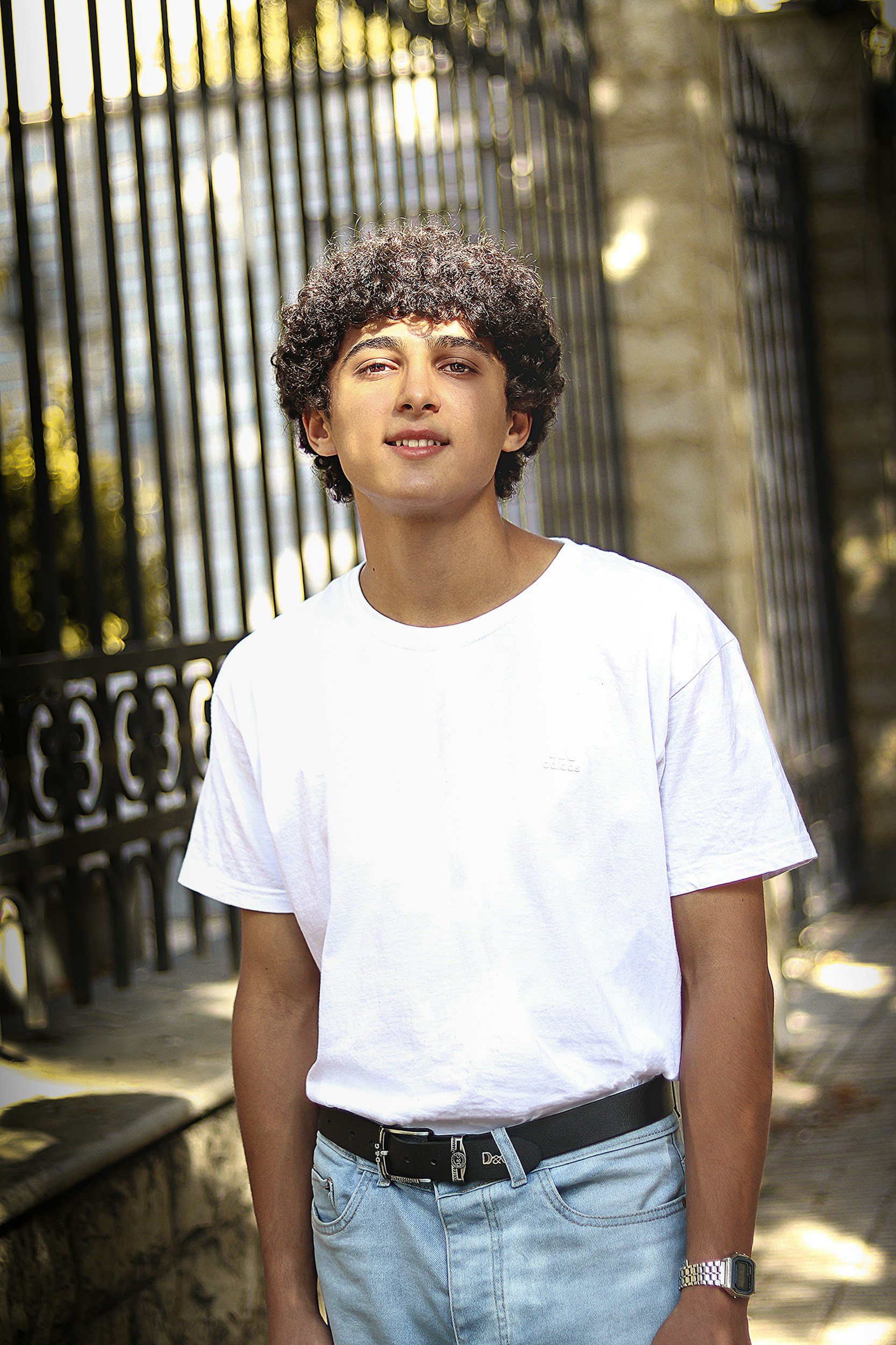 Young man with curly hair wearing a white t-shirt, light blue jeans, and a black belt, standing outdoors near a black wrought iron fence with stone pillars and sunlight filtering through trees.