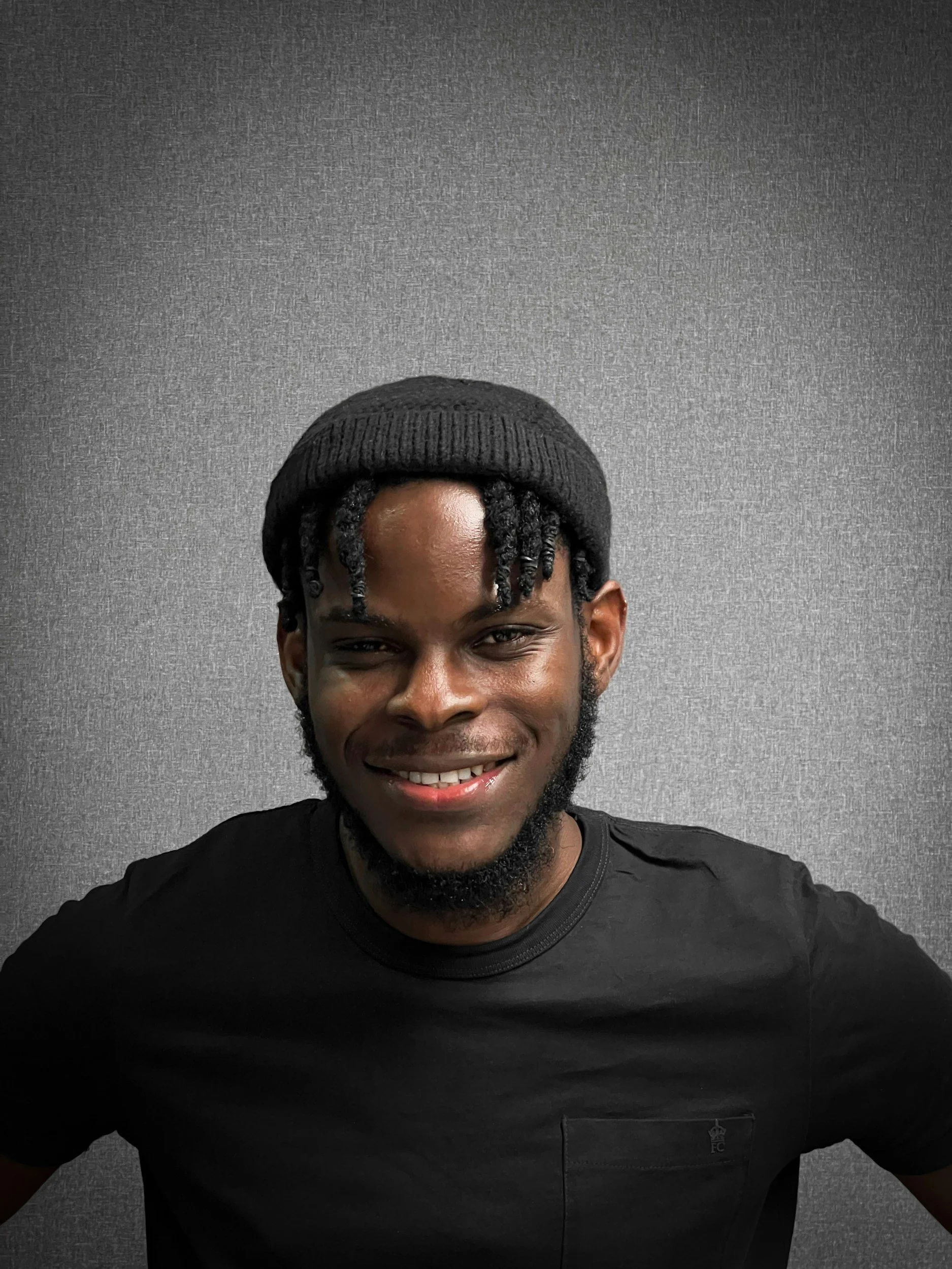 Portrait of a smiling young man wearing a black beanie and black t-shirt, standing against a gray textured background.