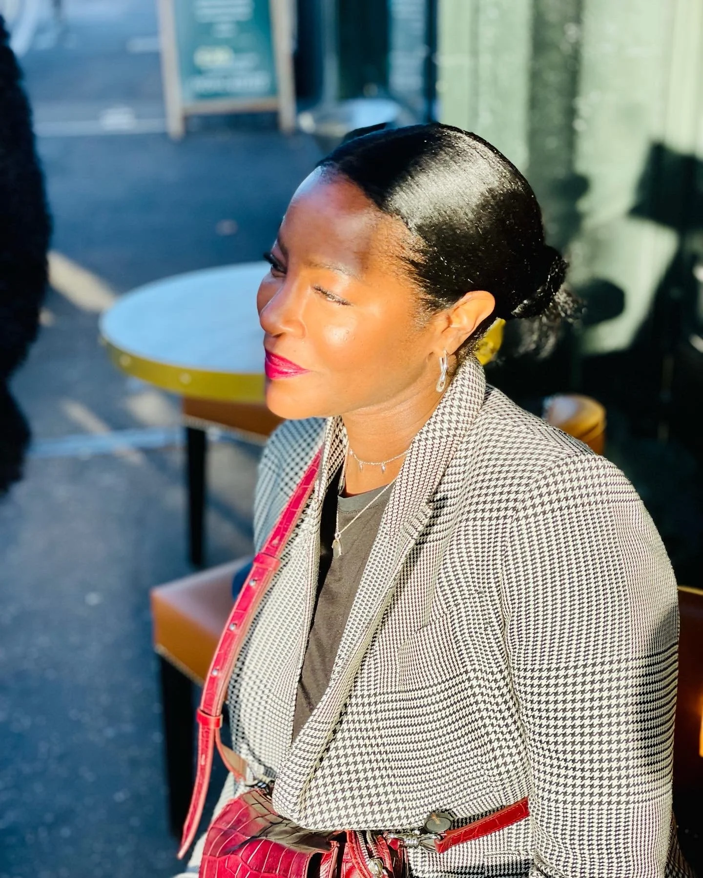 A woman with dark hair styled in a sleek bun, wearing a checkered blazer, red lipstick, and jewelry, sitting outdoor in sunlight.