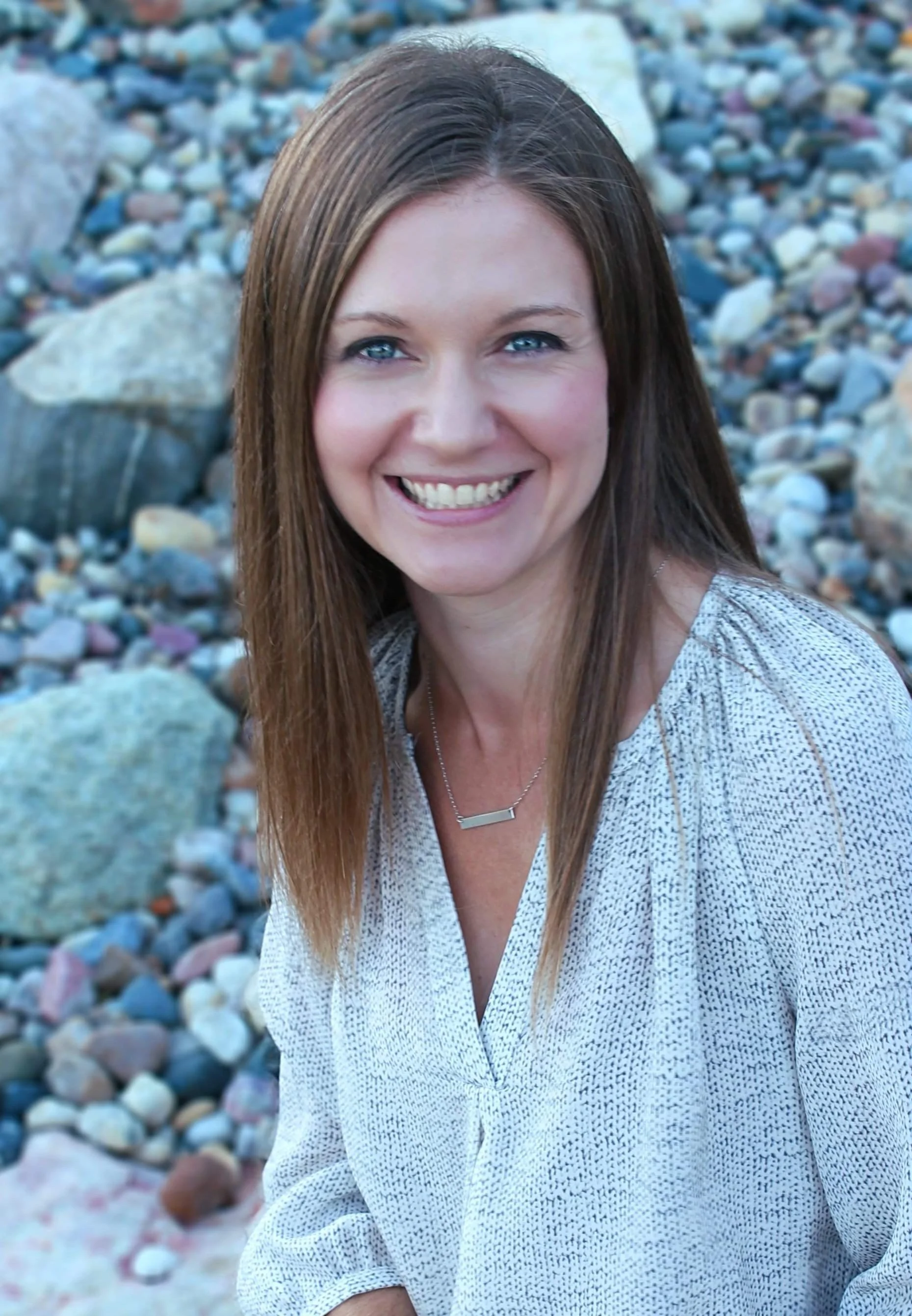 A smiling woman with long brown hair and blue eyes, wearing a light-colored patterned blouse and a silver necklace, standing outdoors on a rocky beach.
