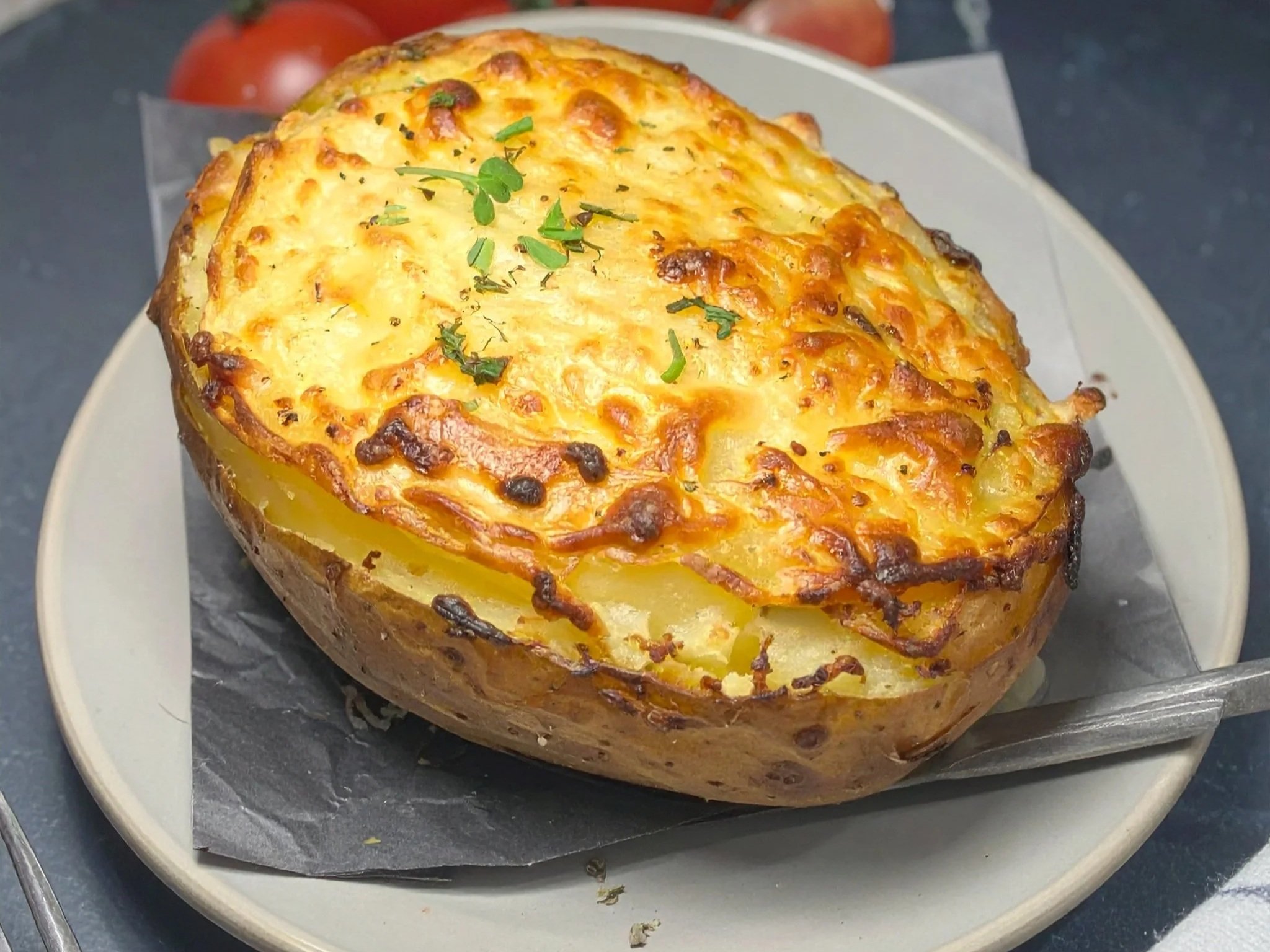 Baked stuffed potato with crispy cheese crust and chopped herbs on top, served on a white plate with black background.