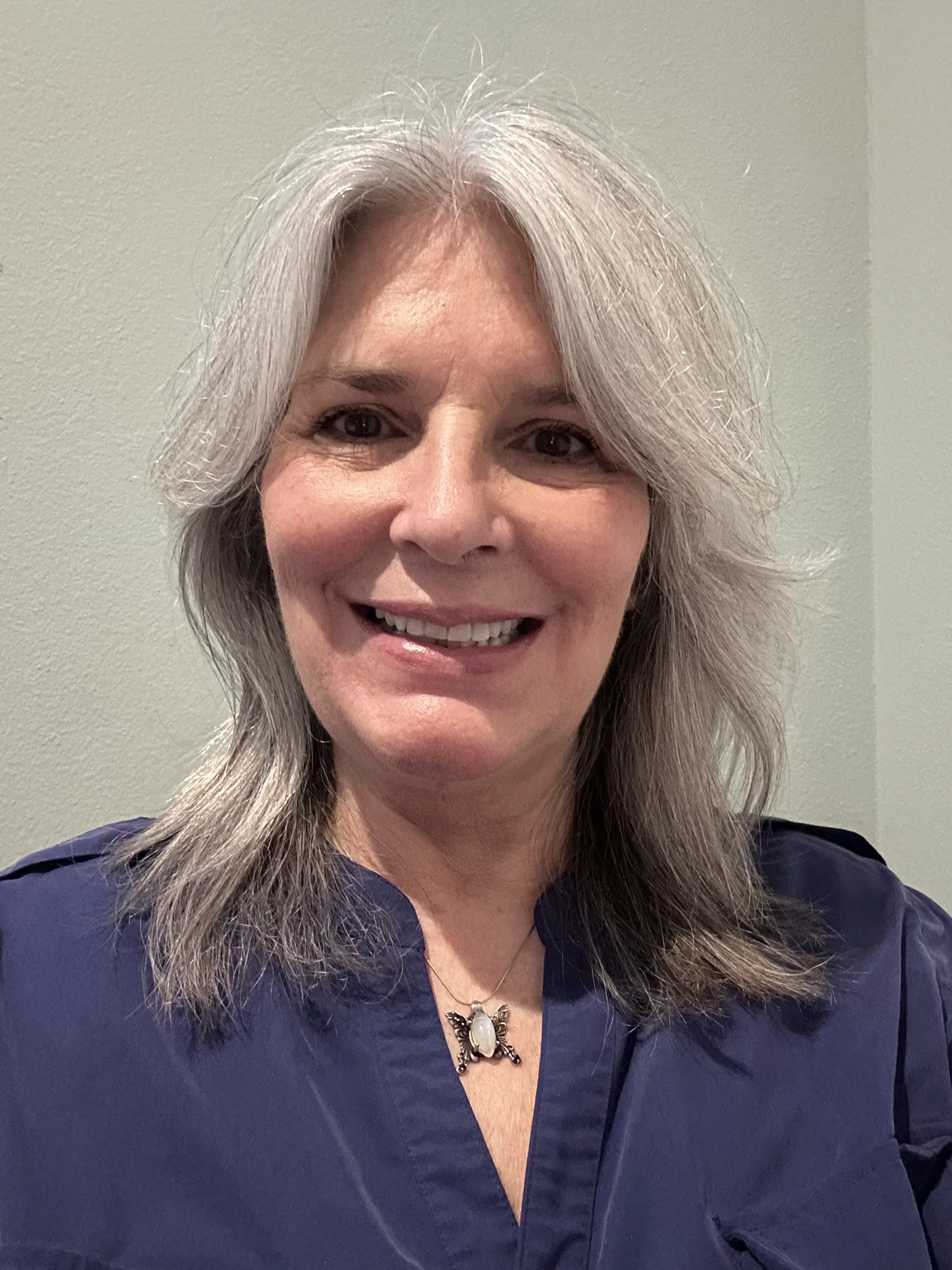 A woman with shoulder-length gray hair smiling, wearing a dark blue top and a necklace with a pendant against a light green wall.