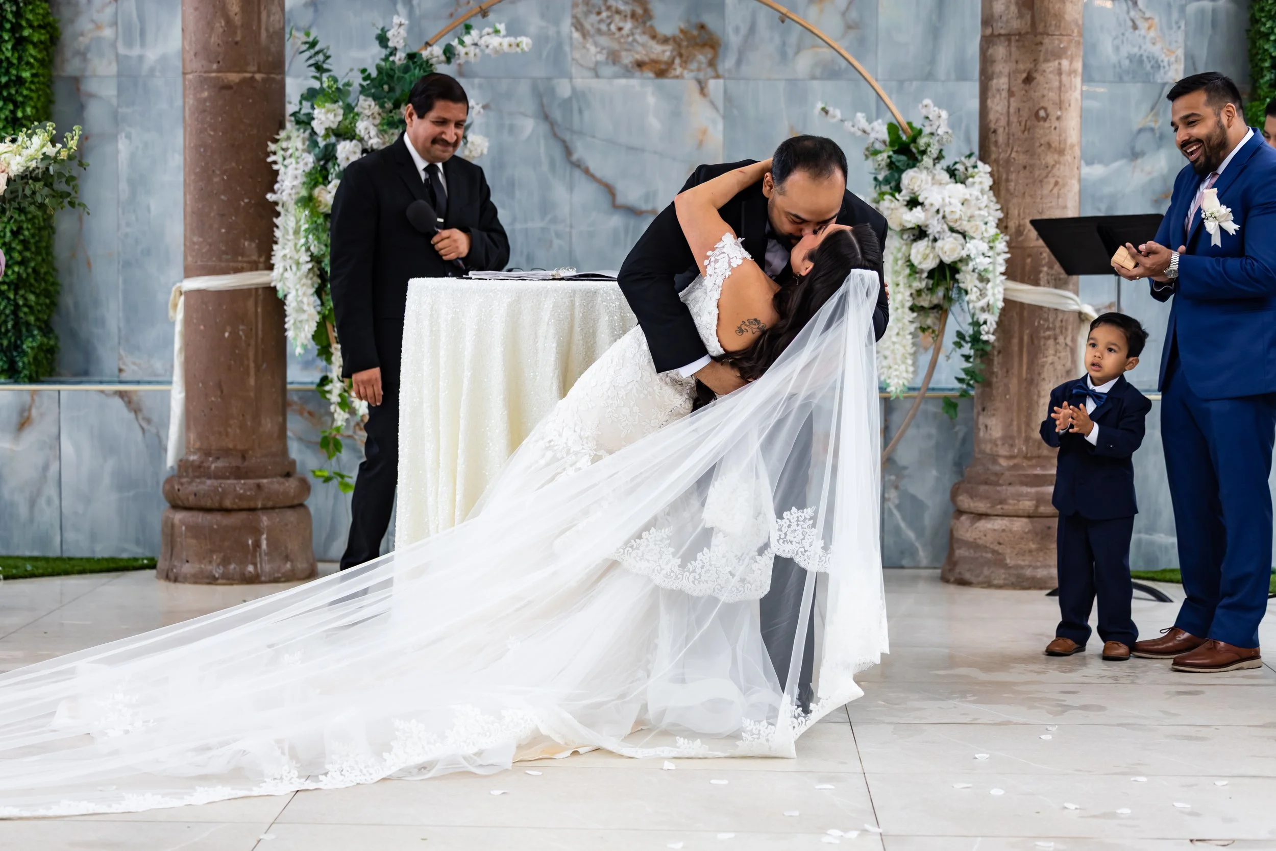 A wedding ceremony where the bride and groom are kissing as they exchange vows, surrounded by wedding officiants and a flower-decorated arch in an indoor setting.