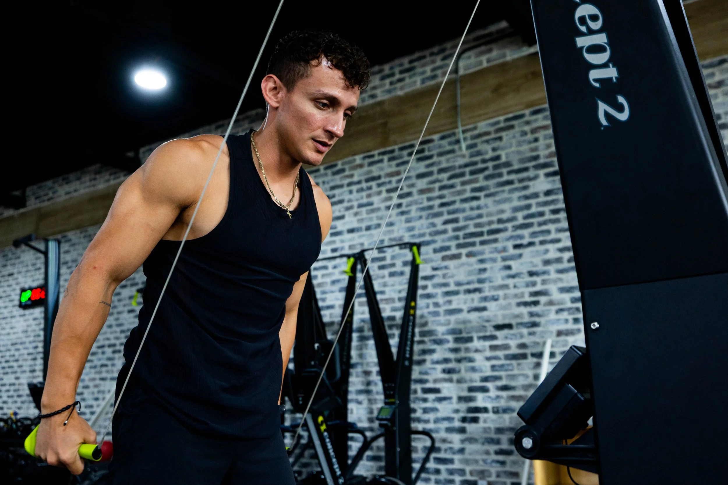 A young man with curly hair and a gold chain is working out in a gym, holding a yellow and black dumbbell, looking at a treadmill or exercise equipment.