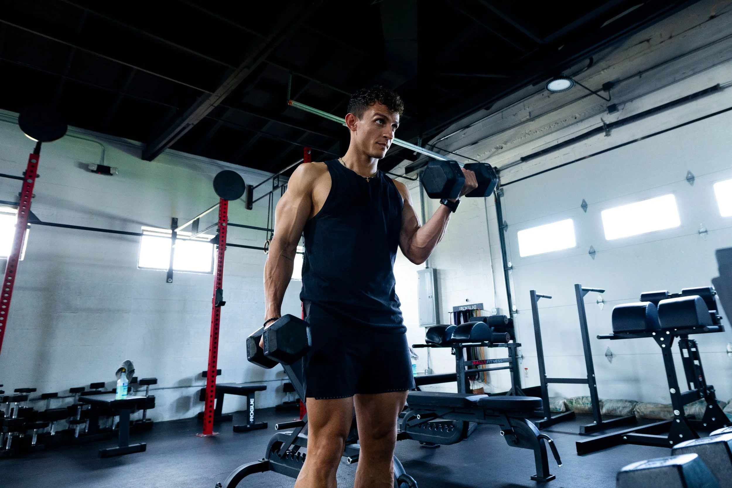 A woman lifting dumbbells in a gym with various equipment like benches, weights, and a squat rack around.