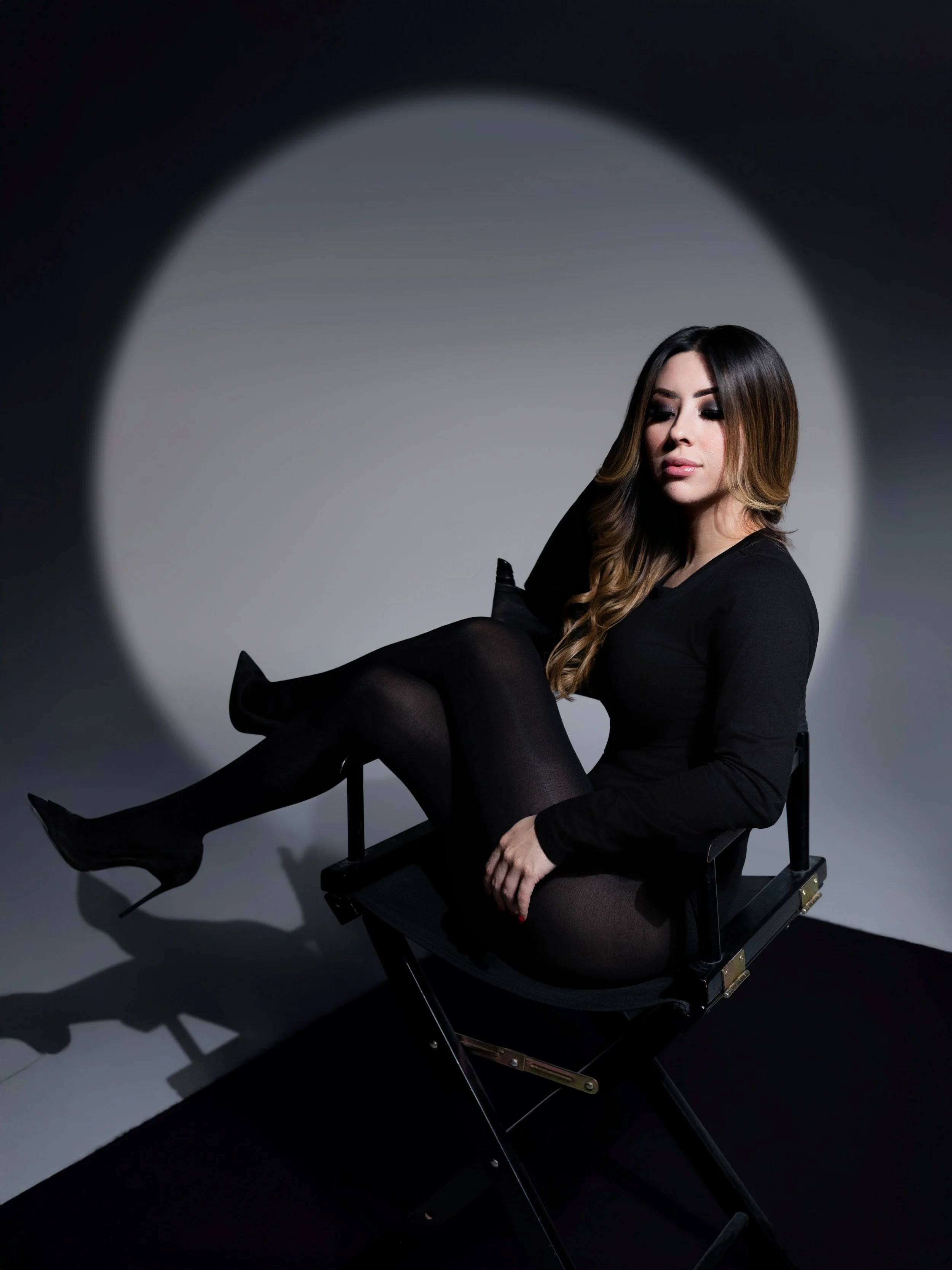 A woman with long wavy hair and dark makeup sitting on a director's chair in a photography studio, with a spotlight creating a circular light on the plain background.