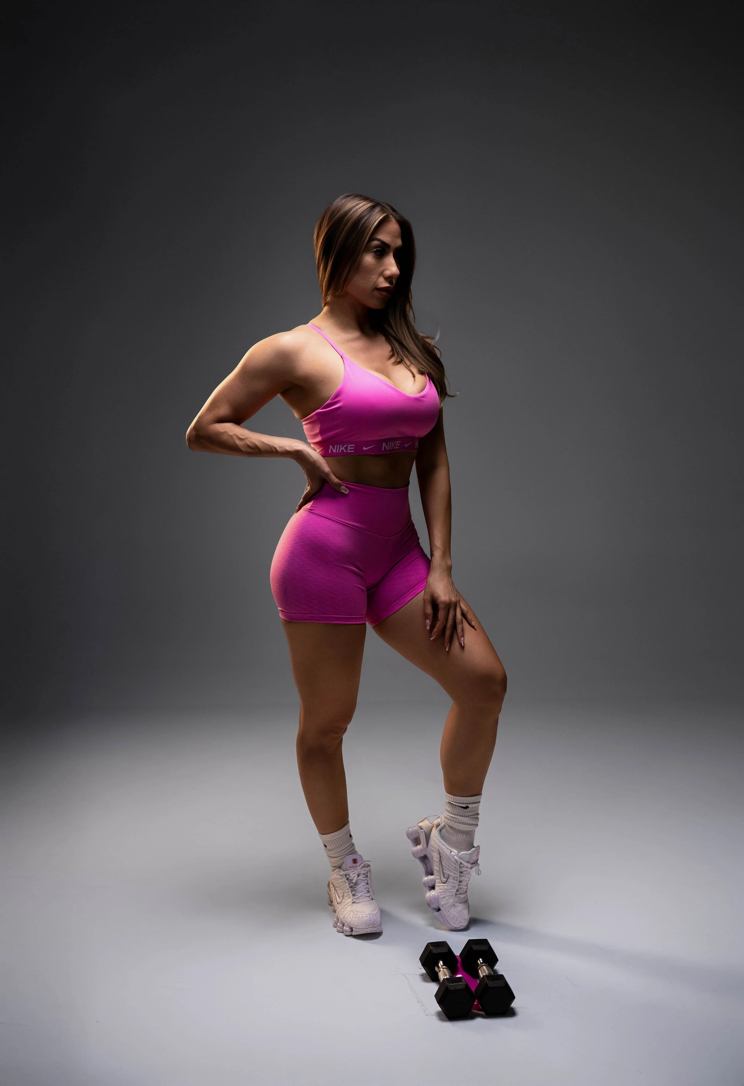 A woman in pink athletic wear, standing in a fitness studio with a gray background, is posing with her hand on her hip next to a black and pink dumbbell and a pair of white athletic shoes and socks.