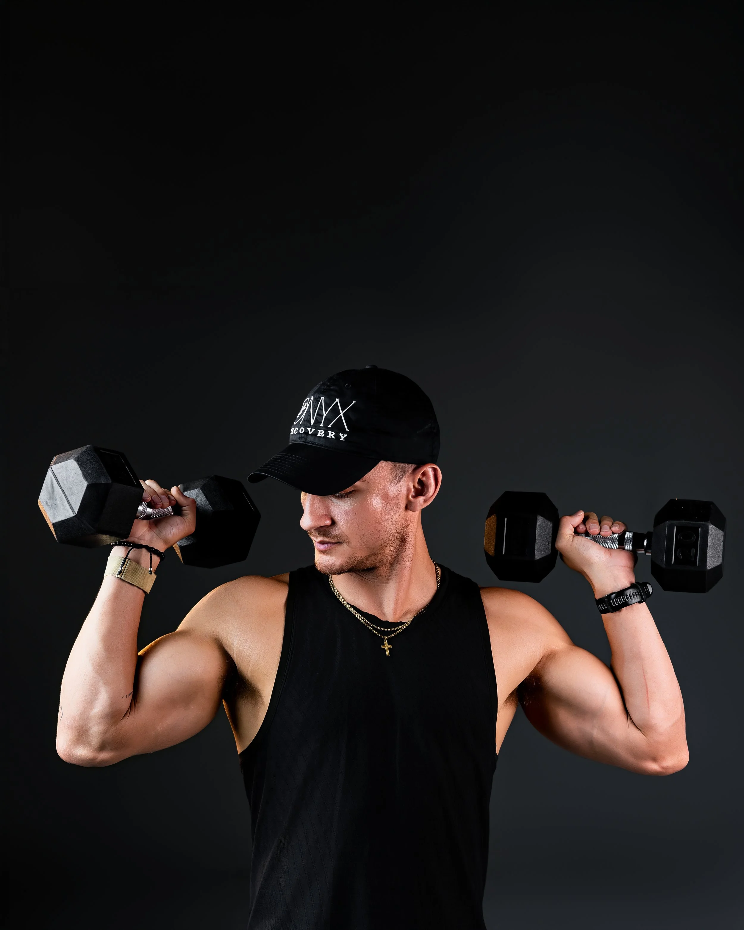 Young man in a black sleeveless shirt lifting dumbbells, wearing a black cap with white text, a wristwatch, a bracelet, and a gold cross necklace, against a dark background.
