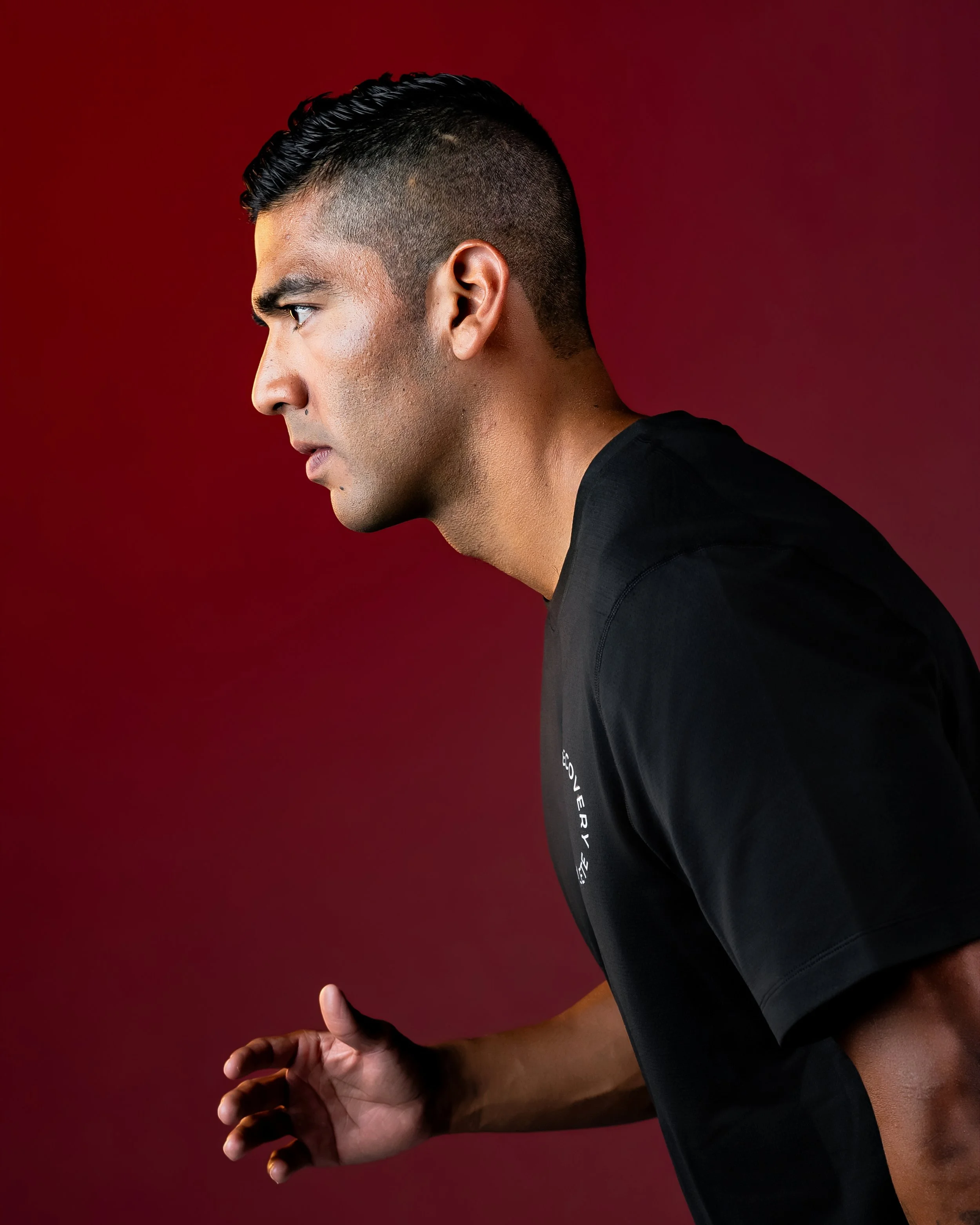 Person in black shirt with short dark hair, holding a large red object behind their head against a deep red background.