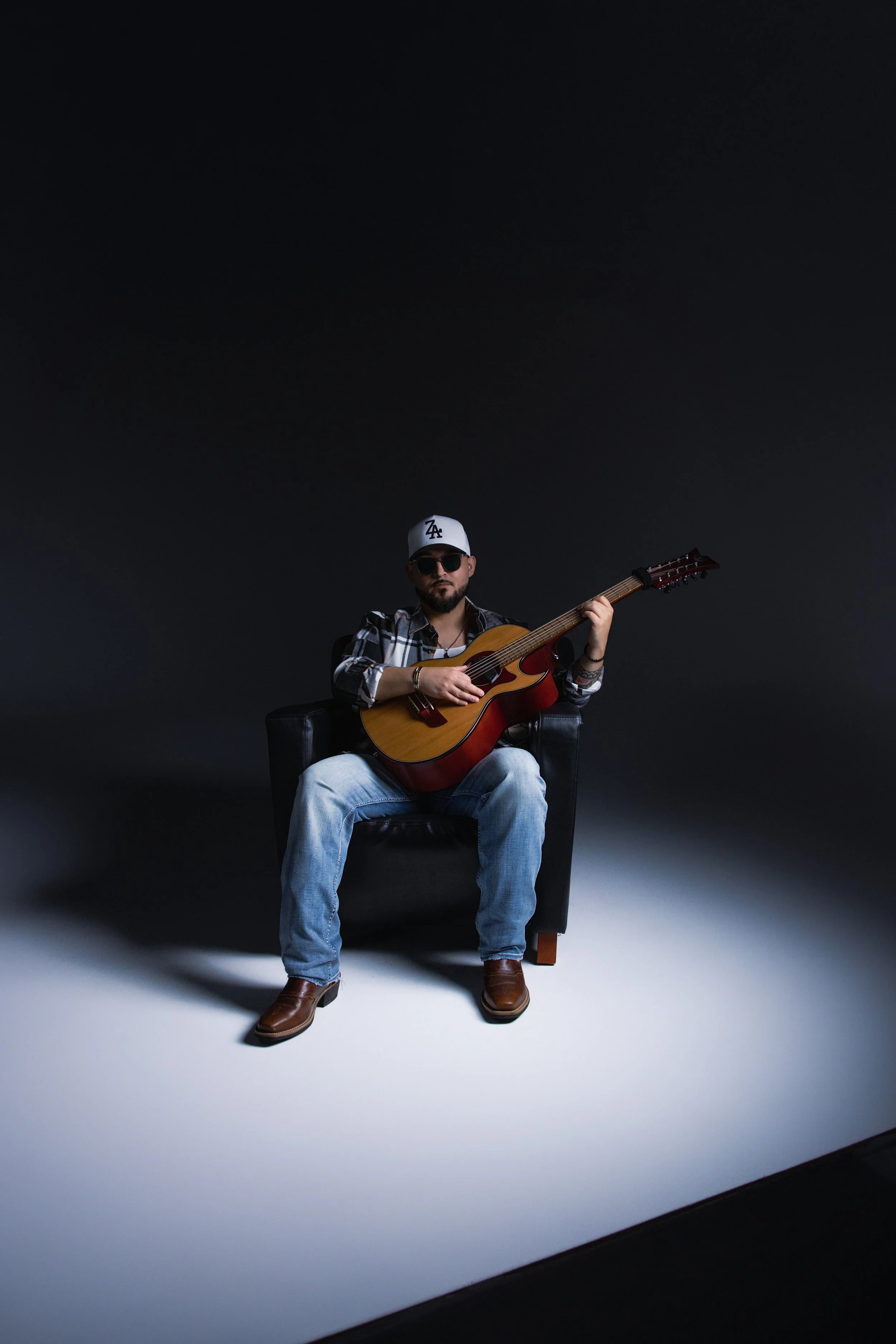 Man sitting on a black chair, wearing sunglasses and a white cap, playing an acoustic guitar against a dark background.
