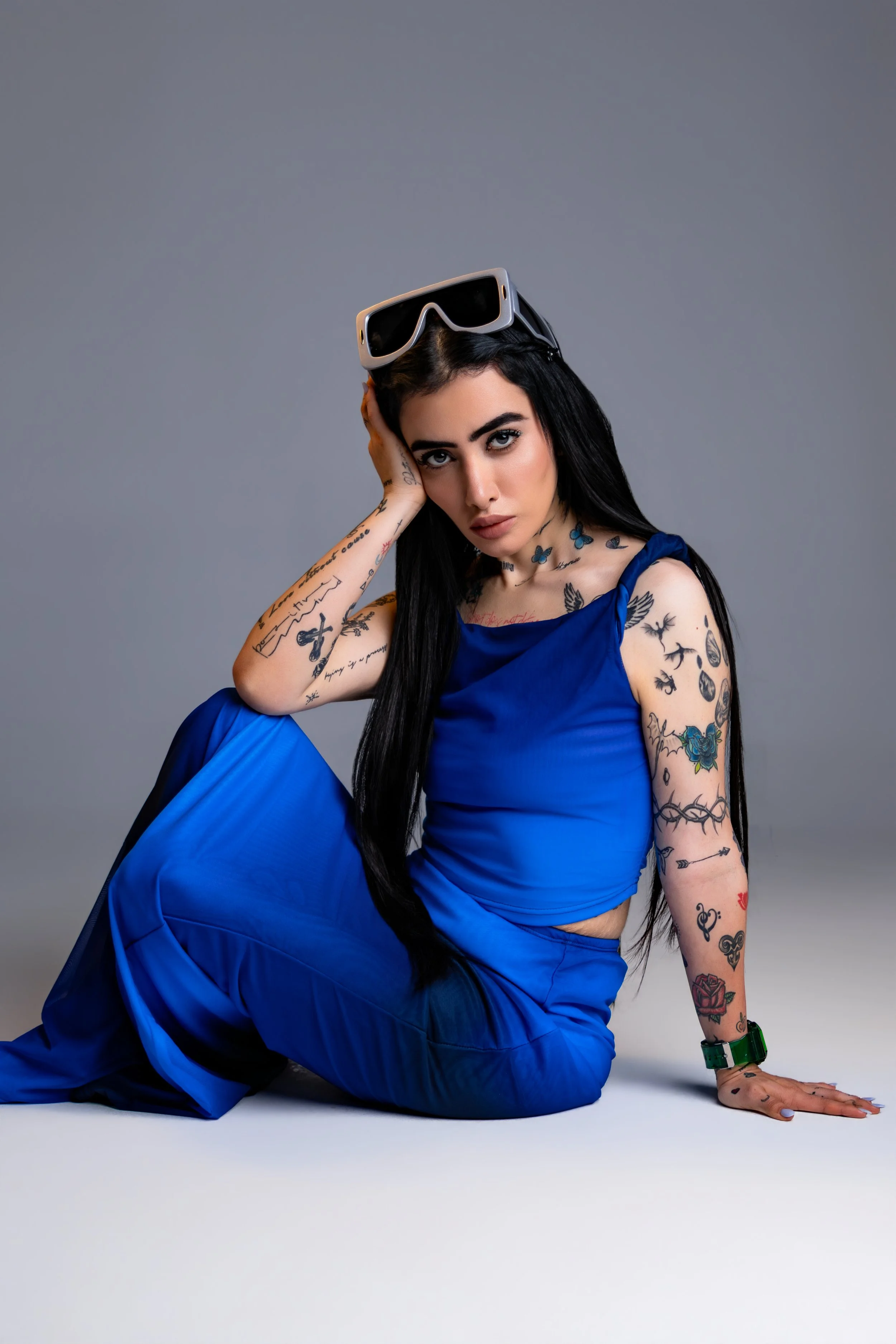 A woman with long black hair, tattoos on her arms and neck, wearing a blue dress and sunglasses on her head, poses sitting on the floor against a gray background.