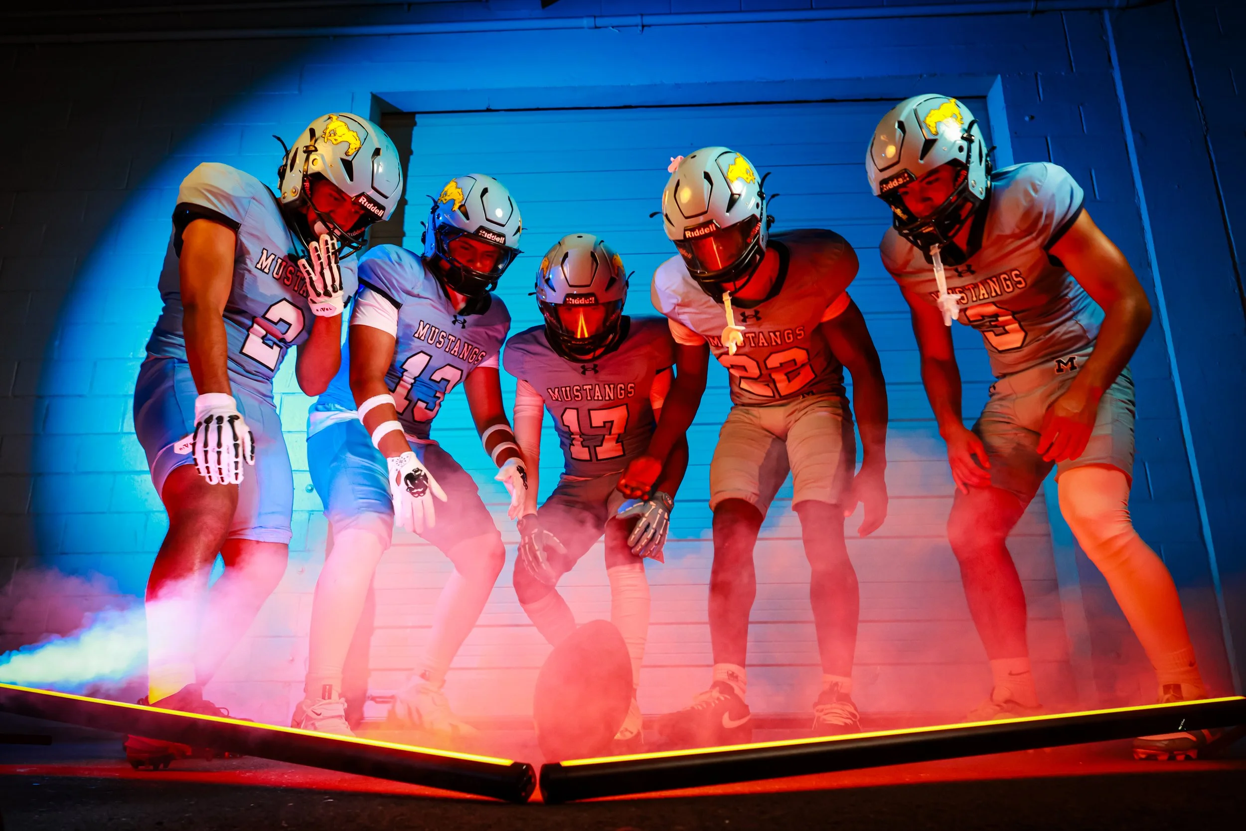 A group of five football players in uniforms and helmets, standing close together in a huddle with mist or smoke at their feet, with a blue-lit background.
