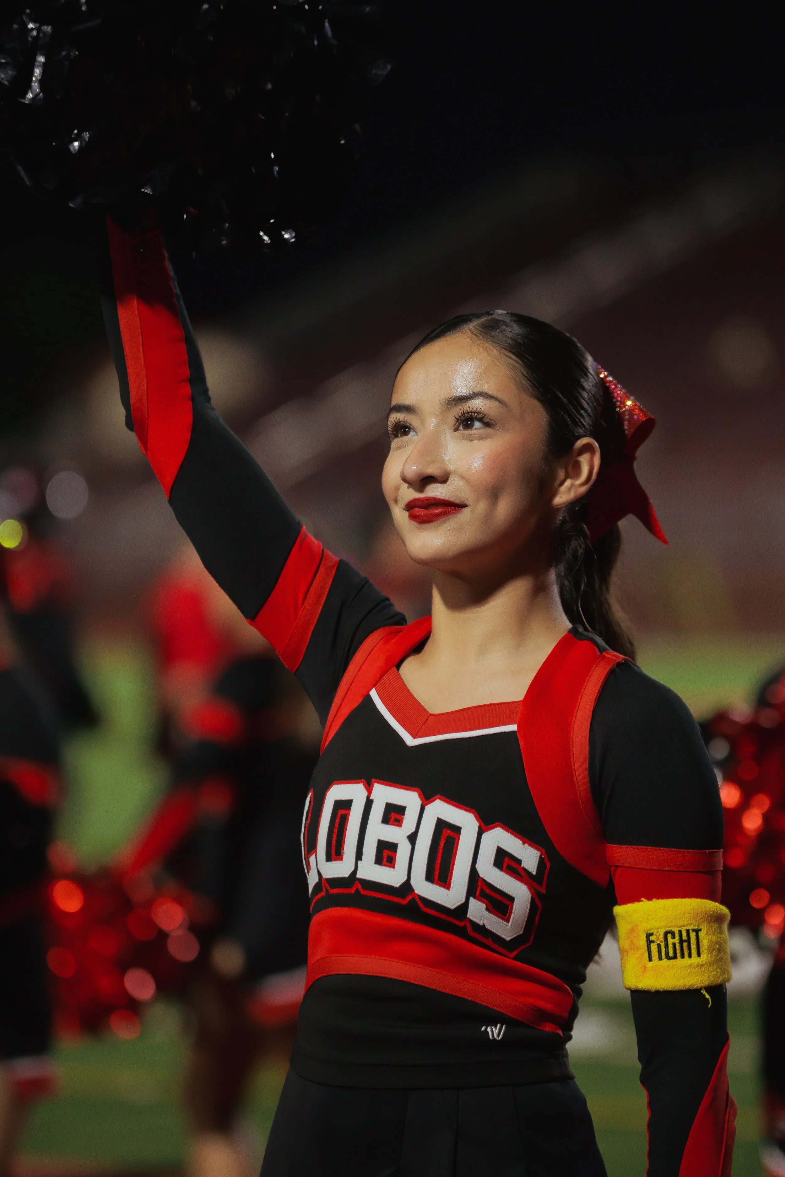 Cheerleader in a black and red cheerleading uniform with the word 'Houston' on the front, raising her arm, and wearing a yellow wristband that says 'Fight' at a sports event with cheerleaders and spectators in the background.