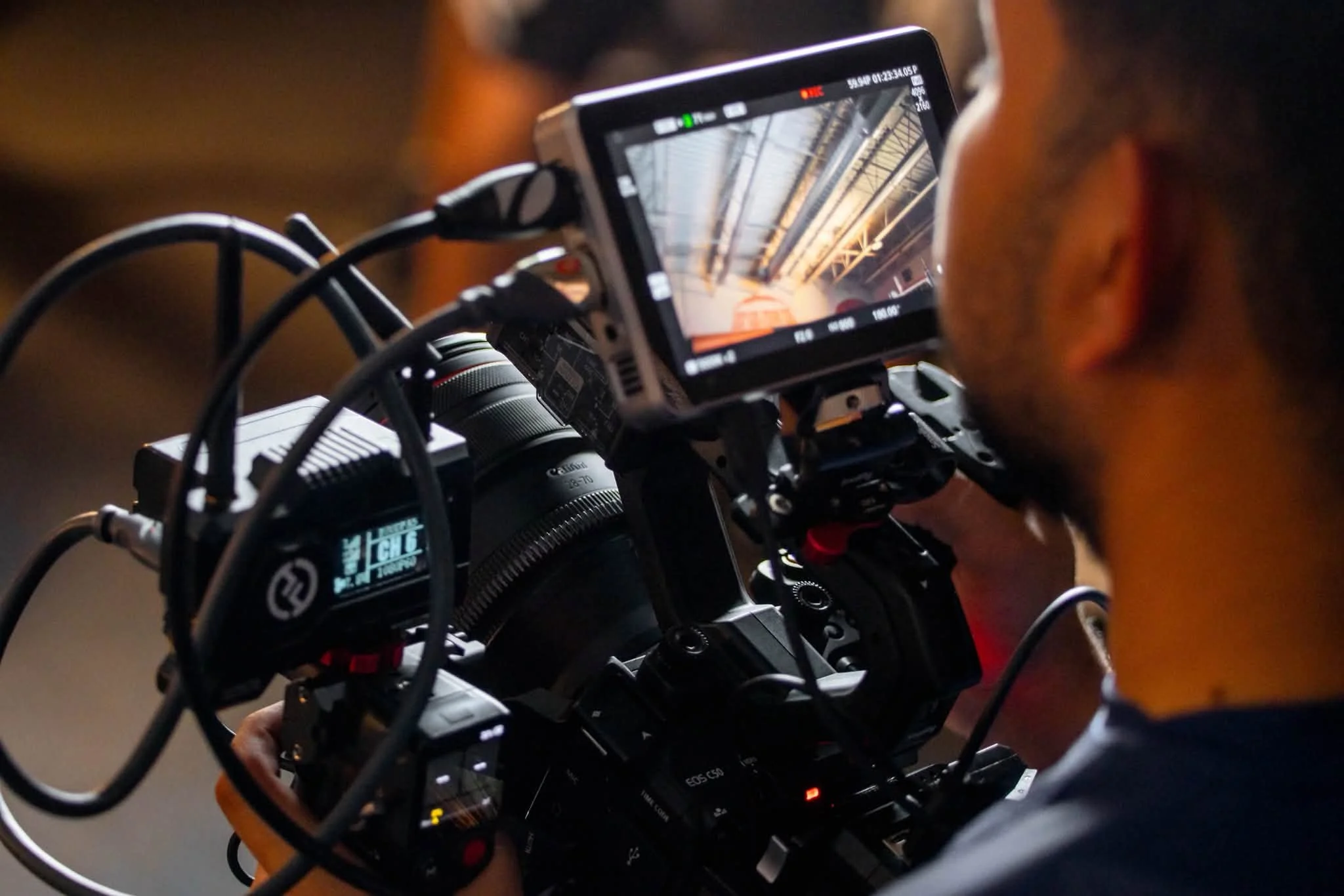 Close-up of a professional video camera on a tripod, with a monitor displaying a shot of an industrial or warehouse interior with high ceilings and beams.
