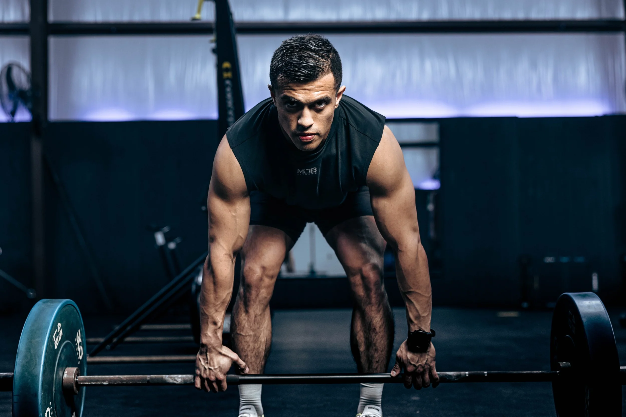 A young man in a black sleeveless shirt and shorts, with short dark hair, is preparing to lift a barbell in a gym. He is crouched down in a starting position, focused and determined, surrounded by gym equipment.