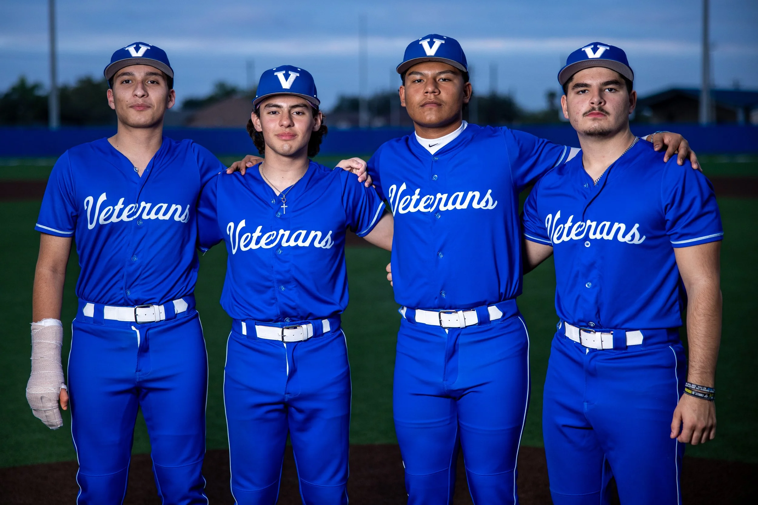 Four young men standing together on a baseball field at dusk, wearing blue baseball uniforms with the word 'Veterans' on the front and blue caps with a white 'V' on the front.