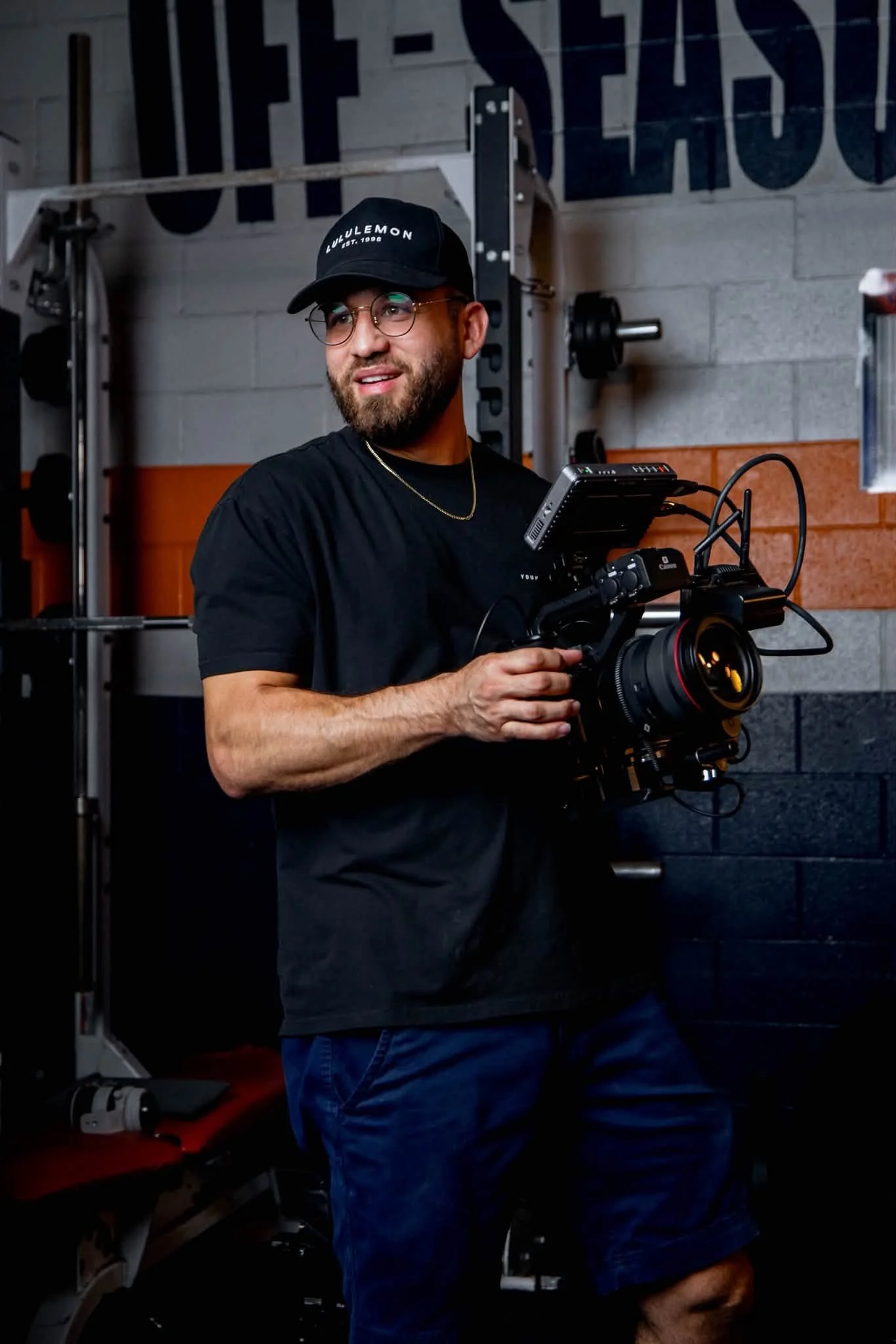 A man wearing glasses, a black T-shirt, a black baseball cap, and a gold chain, holding a professional camera in a gym or workout area with weights and gym equipment in the background.