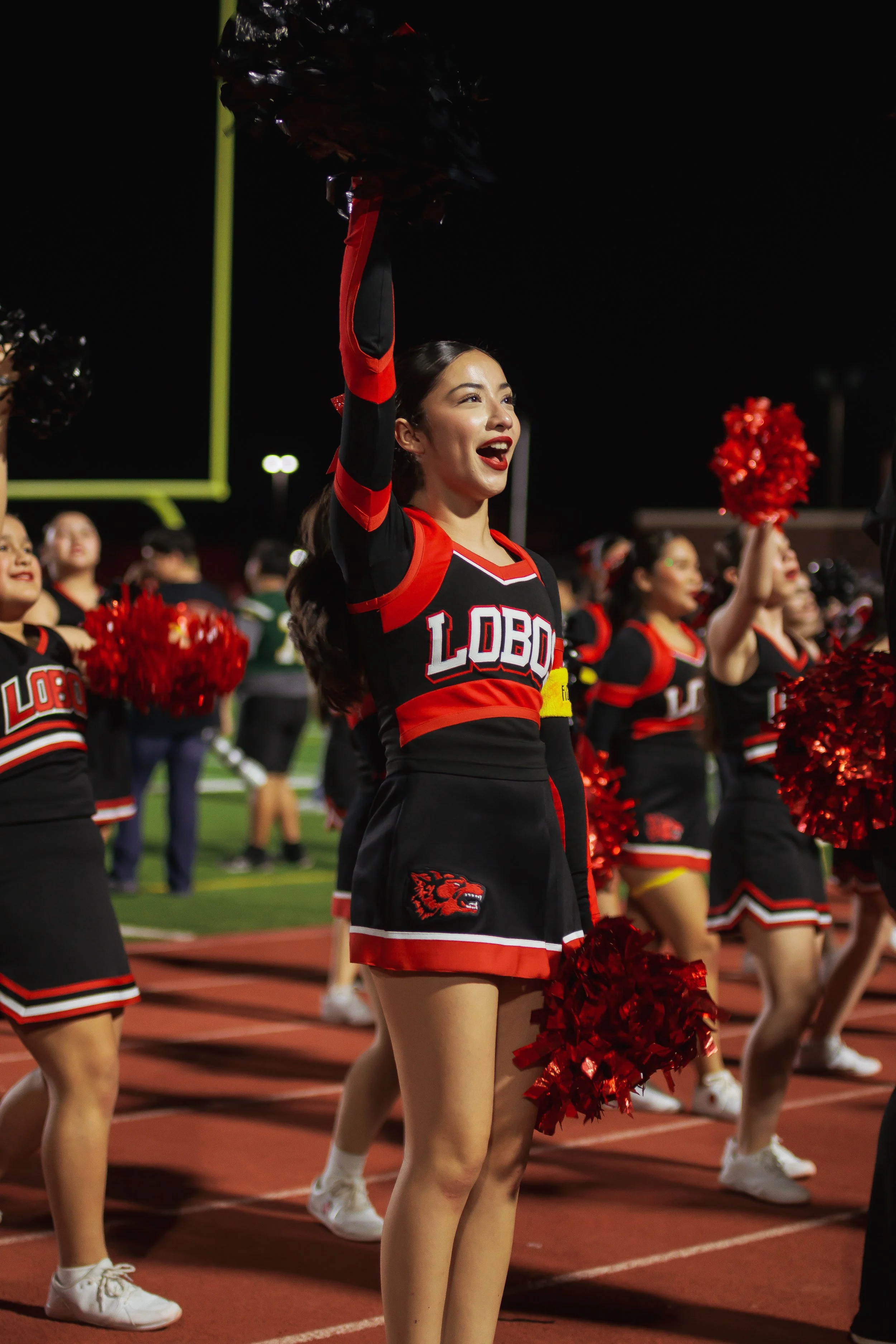 Cheerleaders in black and red uniforms holding red pom-poms during a nighttime football game.