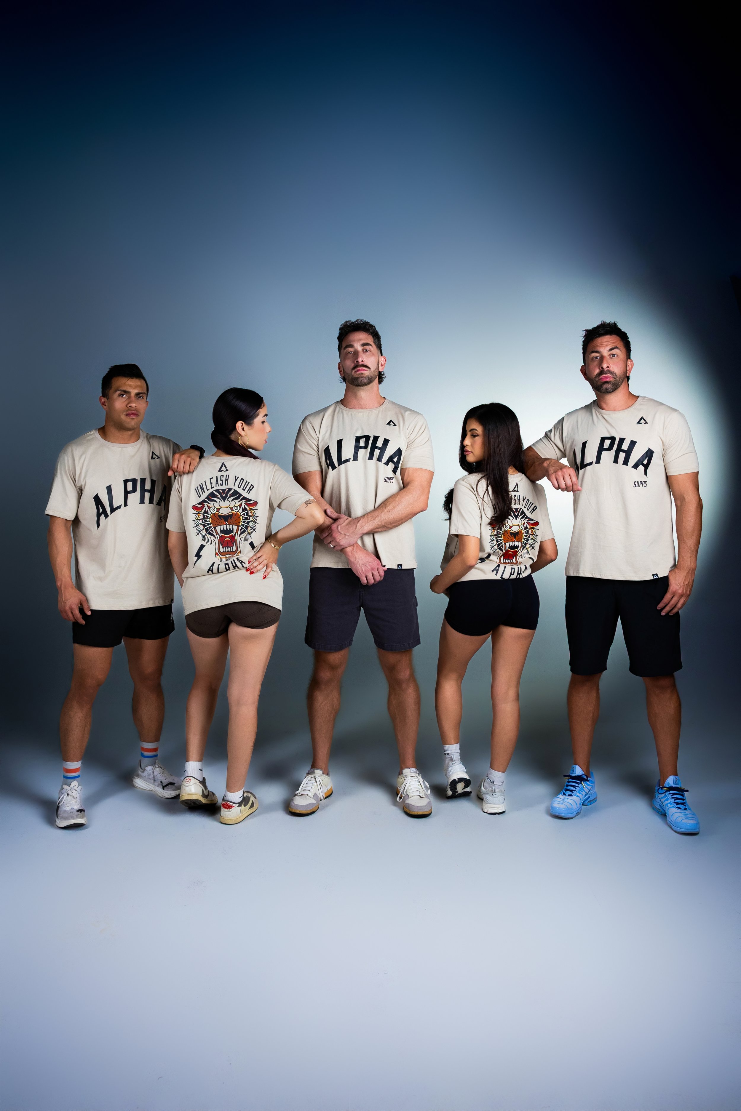 Group of six people standing together, wearing matching cream-colored shirts with the word 'ALPHA' and a tiger graphic, posing confidently in a studio with a plain background.