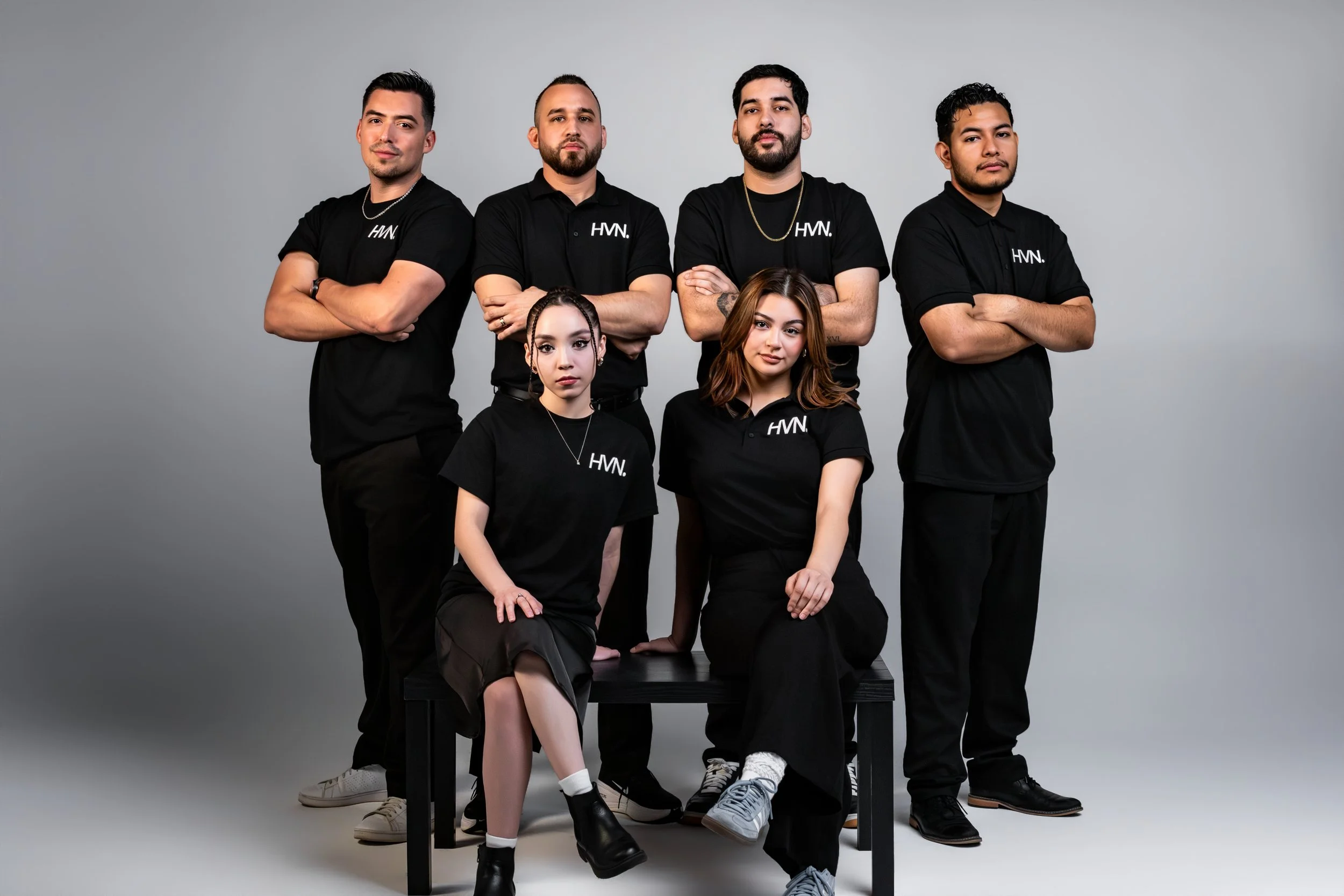 Group of seven diverse young adults posing for a team or promotional photo against a plain gray background. Four men standing in the back row and three women seated in front. They are dressed in black shirts, some with logos, with confident expressions and cross-armed or relaxed poses.