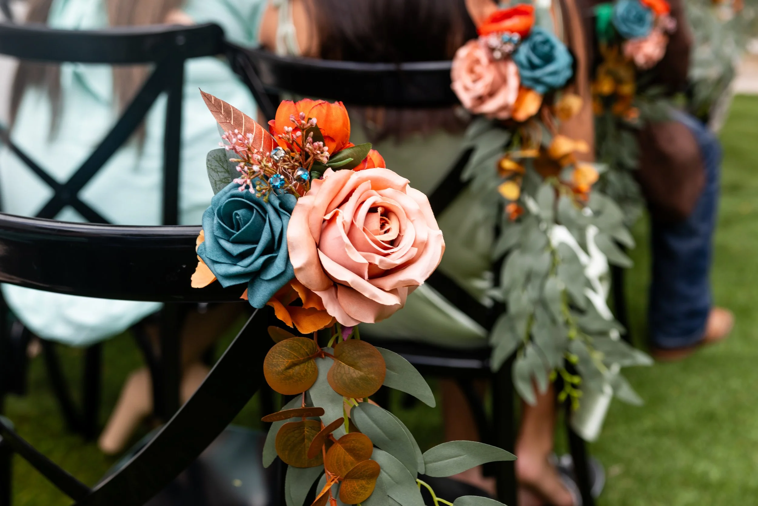 Close-up of decorative flowers on a black metal chair, including pink and orange roses, blue fabric flowers, and various green foliage, with a blurred background of green grass and a person in a plaid shirt.