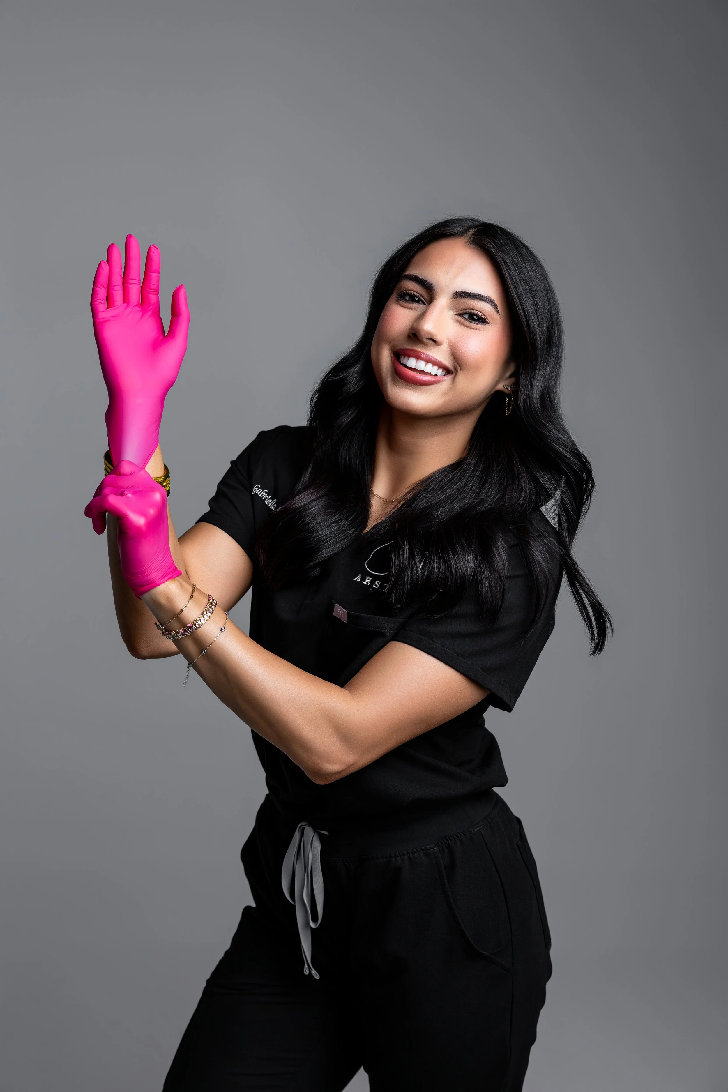 A young woman with long black hair and a bright smile, wearing a black scrub top and black pants, holds up her right arm covered in a pink rubber glove, waving at the camera against a plain grey background.