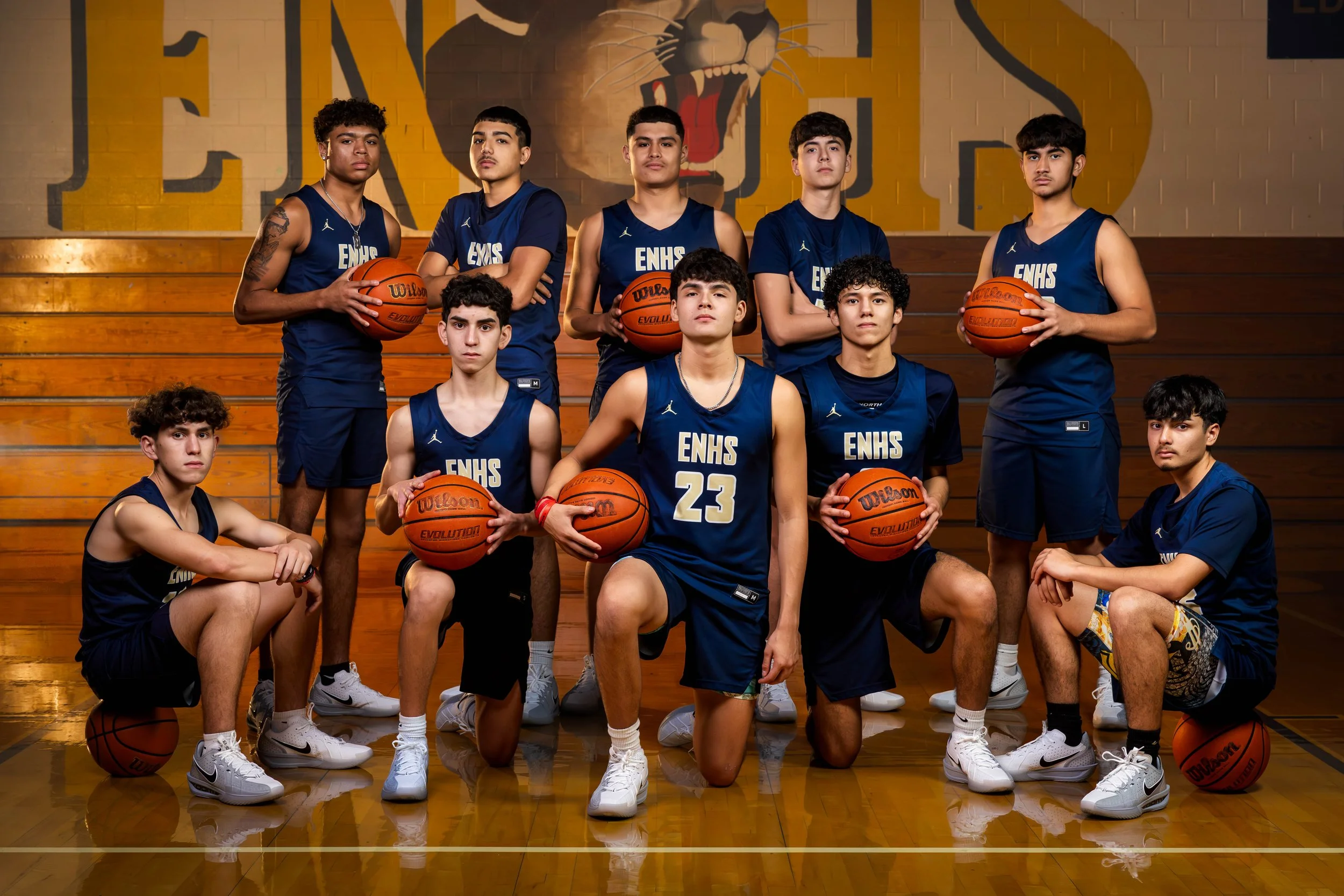 A group of young male basketball players in blue jerseys with 'ENHS' on them, posing for a team photo in a gymnasium. Some are standing while others are kneeling or sitting, holding basketballs. The background features a large painted lion face on th