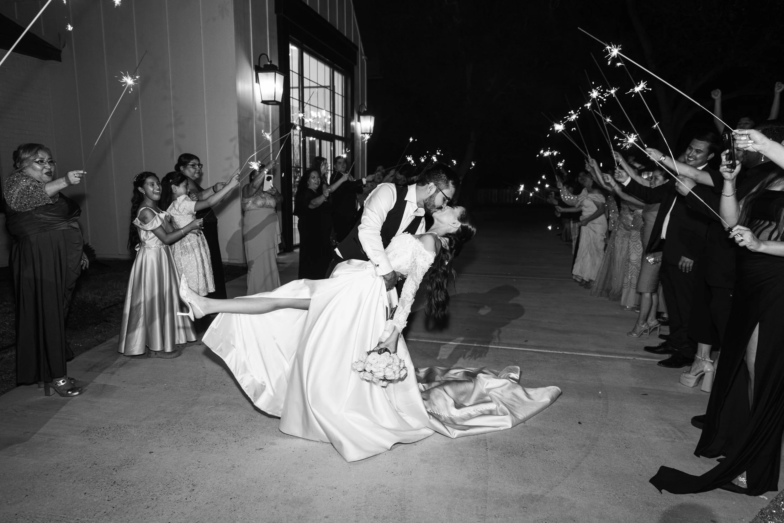 A couple in wedding attire kissing while being dipped by each other during a celebration with guests holding sparklers.