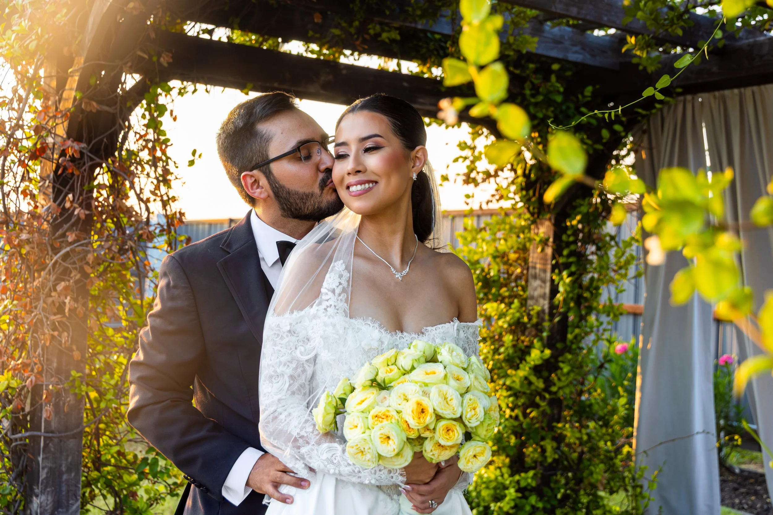 A newlywed couple, the groom kissing the bride's temple, holding a bouquet of yellow roses, standing outdoors amidst greenery and sunlight.