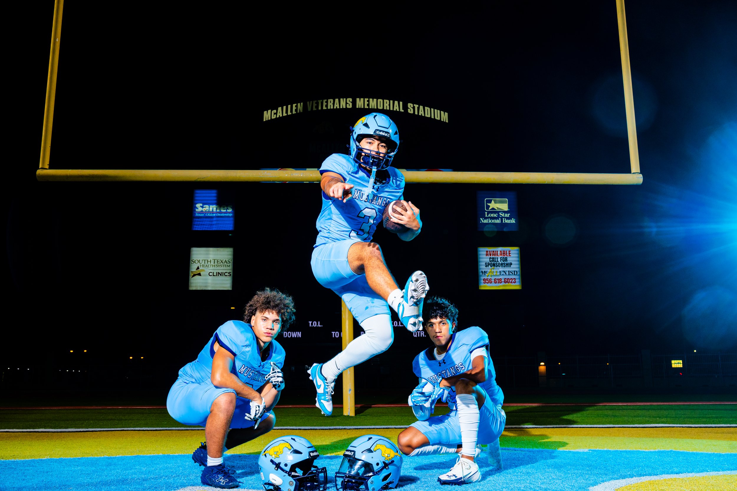 A group of high school football players in blue uniforms, with helmets placed on the ground, posing on a football field at night under stadium lights.