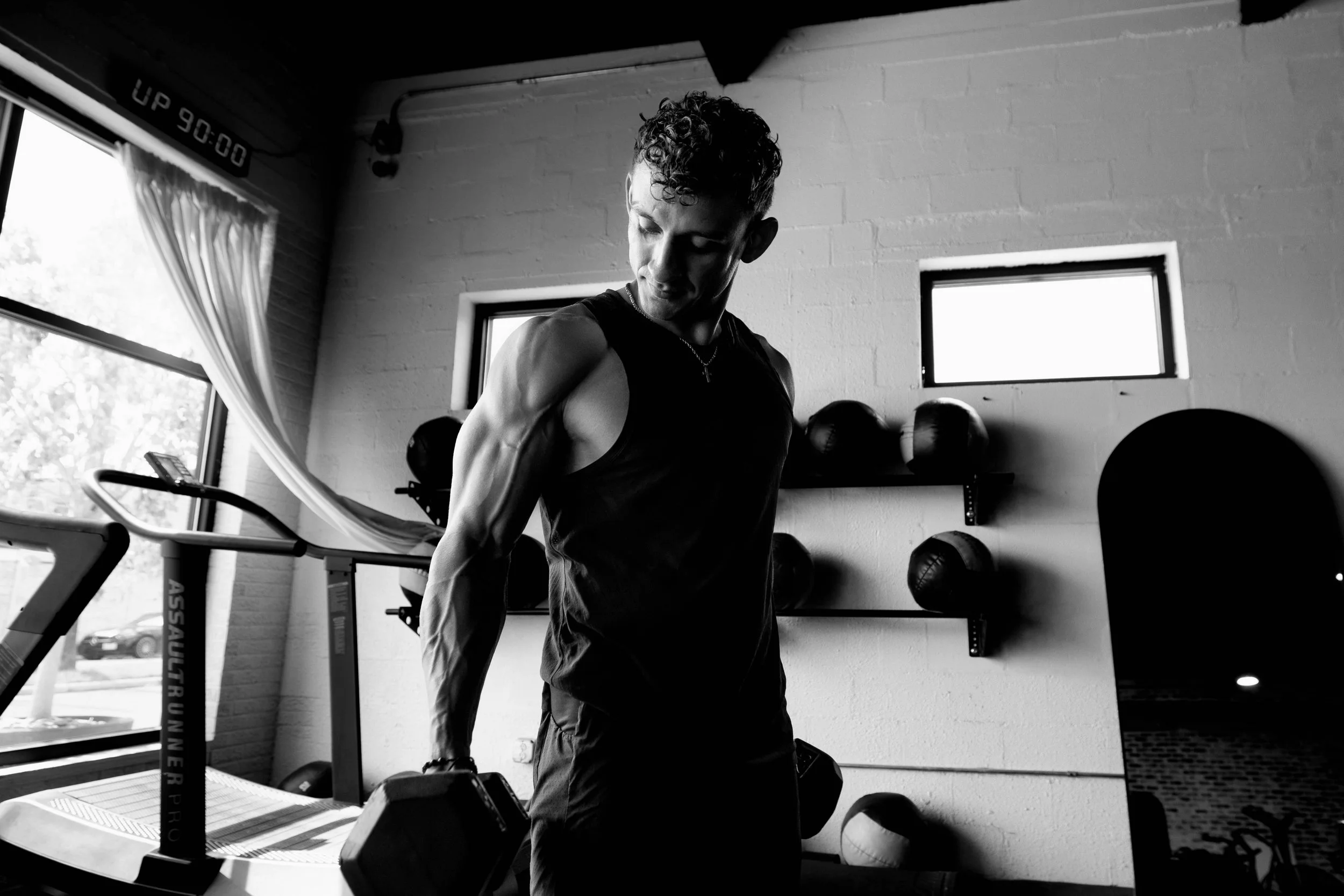 A man with curly hair and a sleeveless shirt lifting a dumbbell in a gym, with gym equipment and windows in the background.