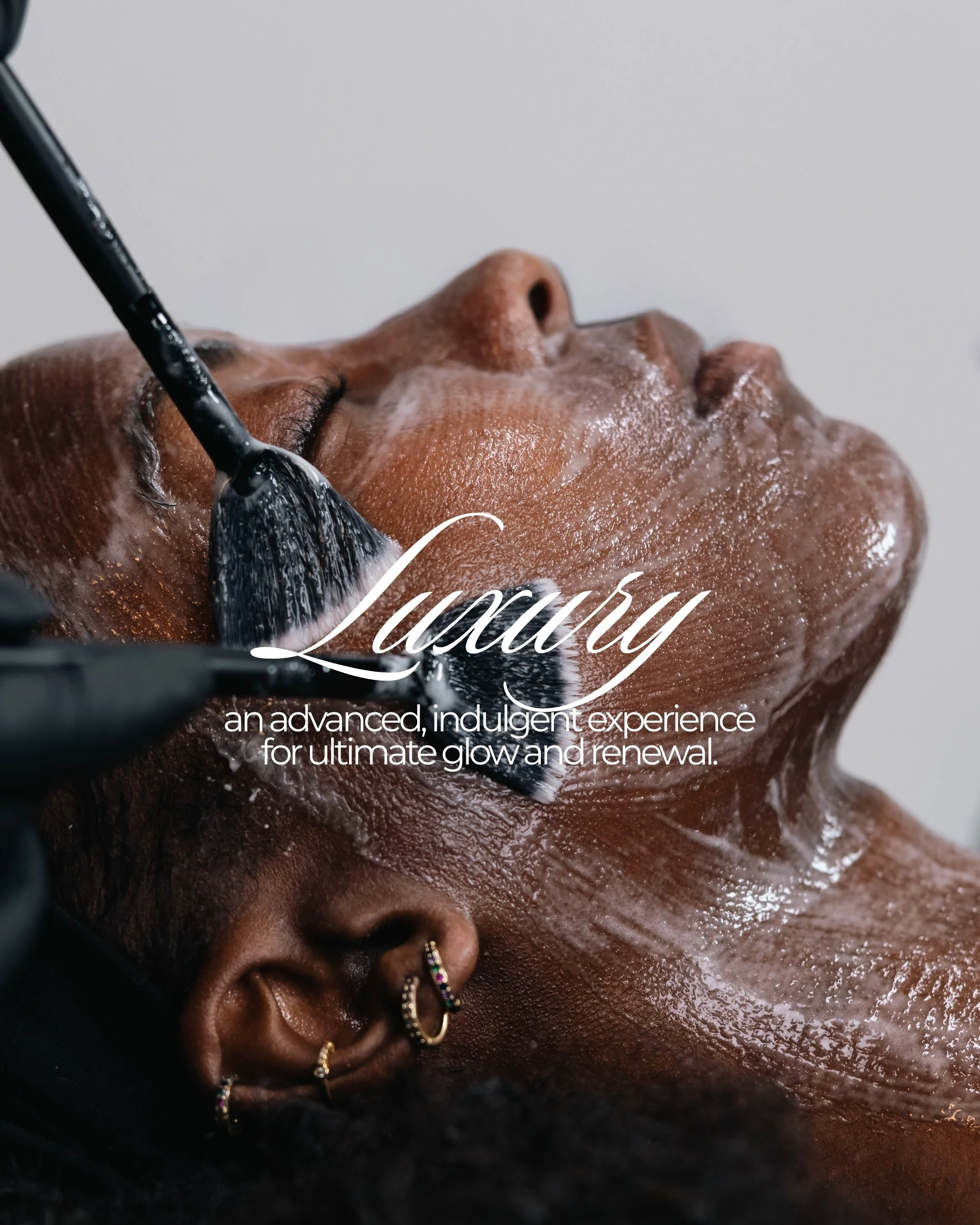Close-up of a person with dark skin receiving a facial treatment, with soap or cleansing foam applied to their face, and someone using a black applicator brush to apply or remove it.
