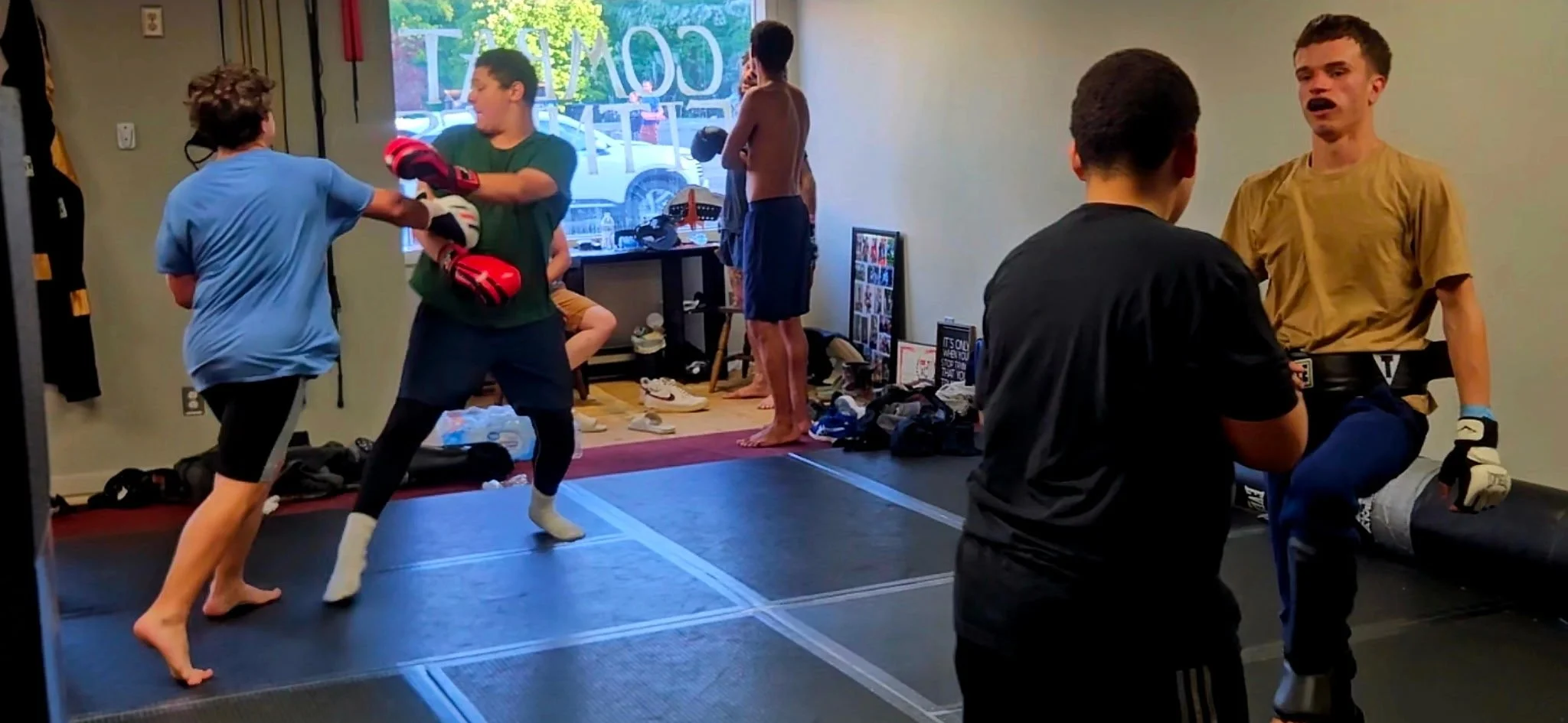 Kids practicing martial arts or kickboxing in a room, with some holding gloves and others sparring or observing, surrounded by personal belongings and a large window in the background.
