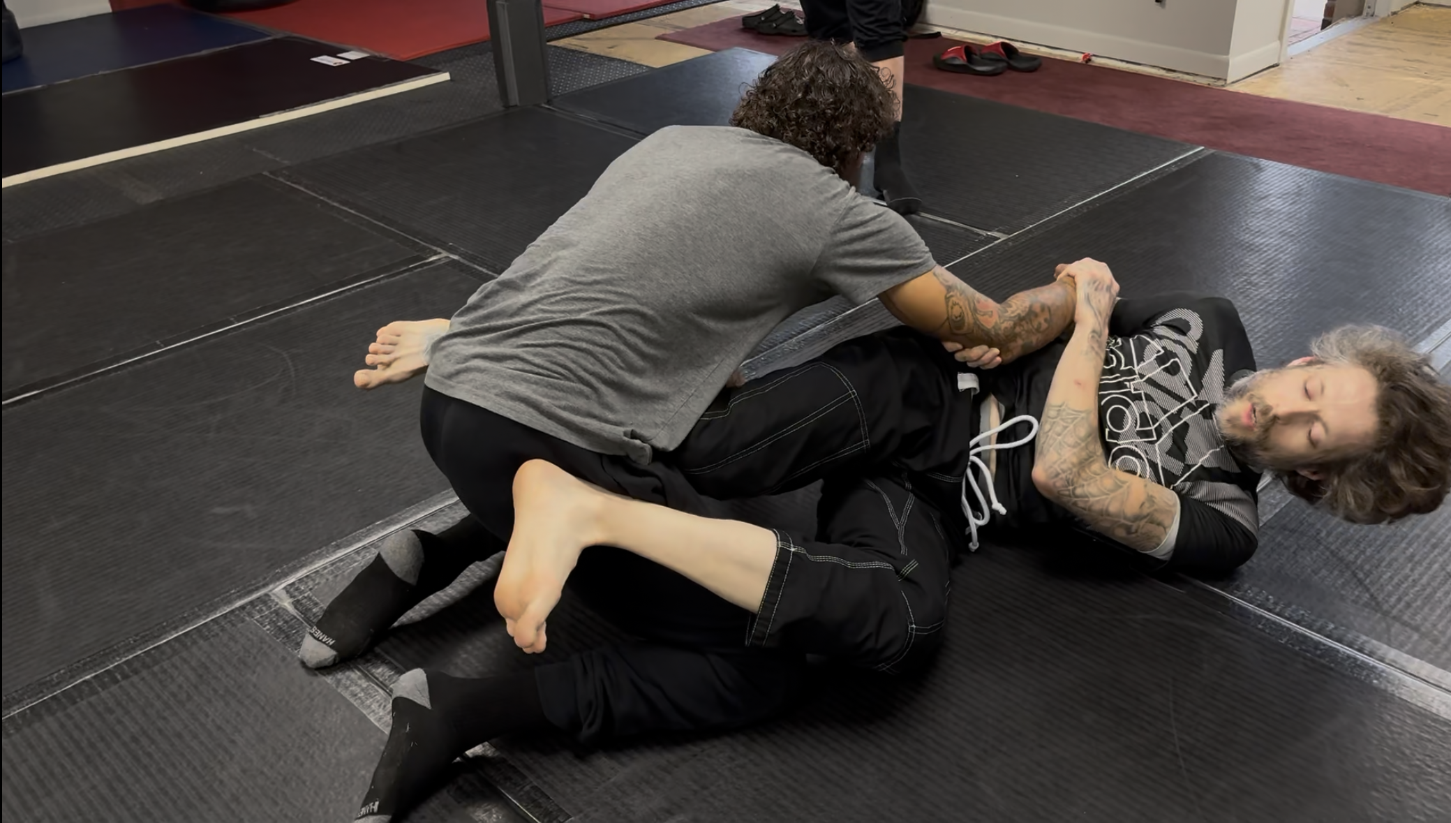 Two men engaged in a Brazilian jiu-jitsu training session on black mats in a gym. One man is on his side, the other is on top, both wearing athletic clothing. The man on top has curly hair and tattoos on his arms, while the man below has tanned skin and short hair.