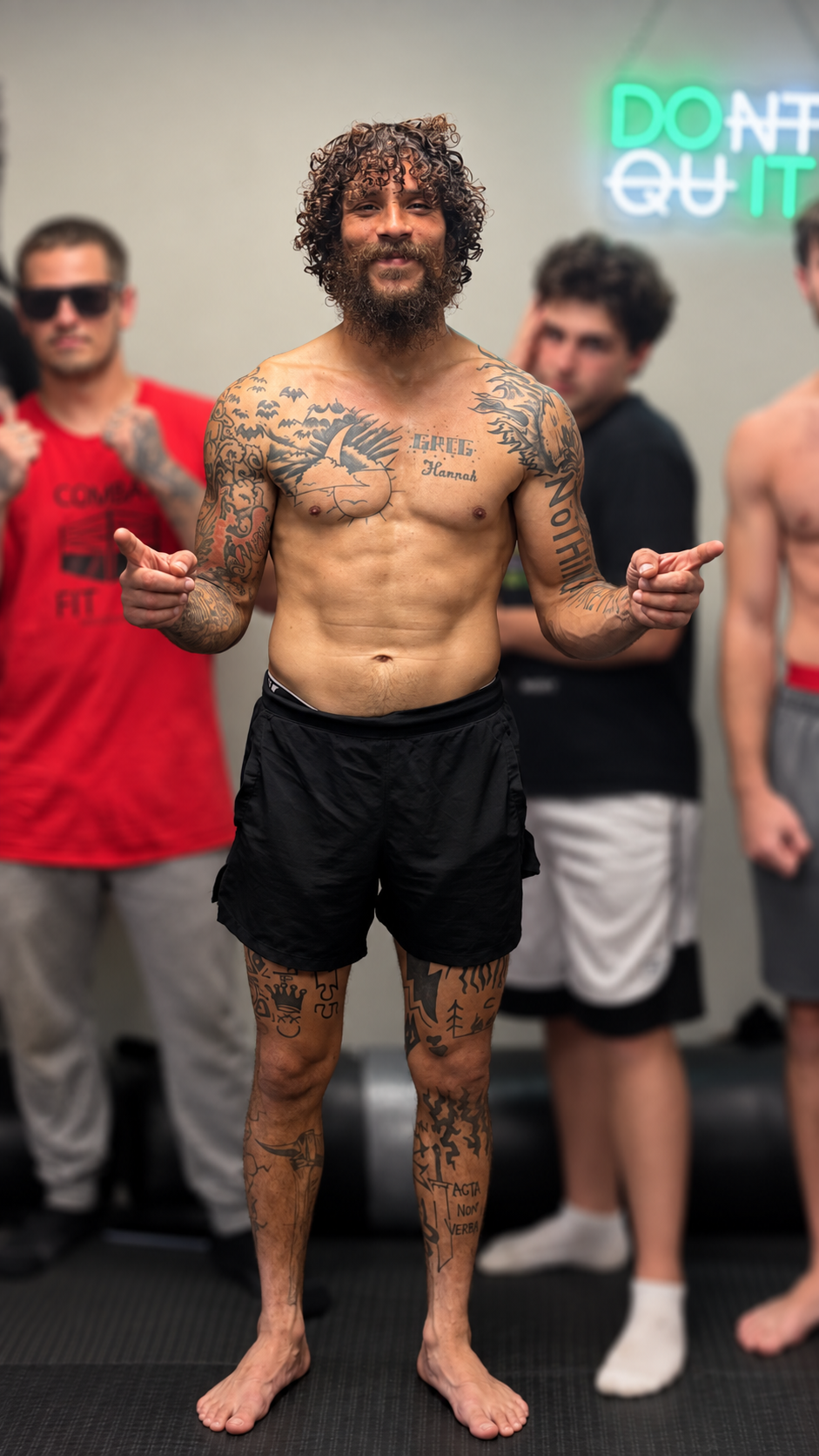 A tattooed man smiling and pointing with both hands, standing barefoot in front of a group of people in an indoor setting, with a neon sign in the background.