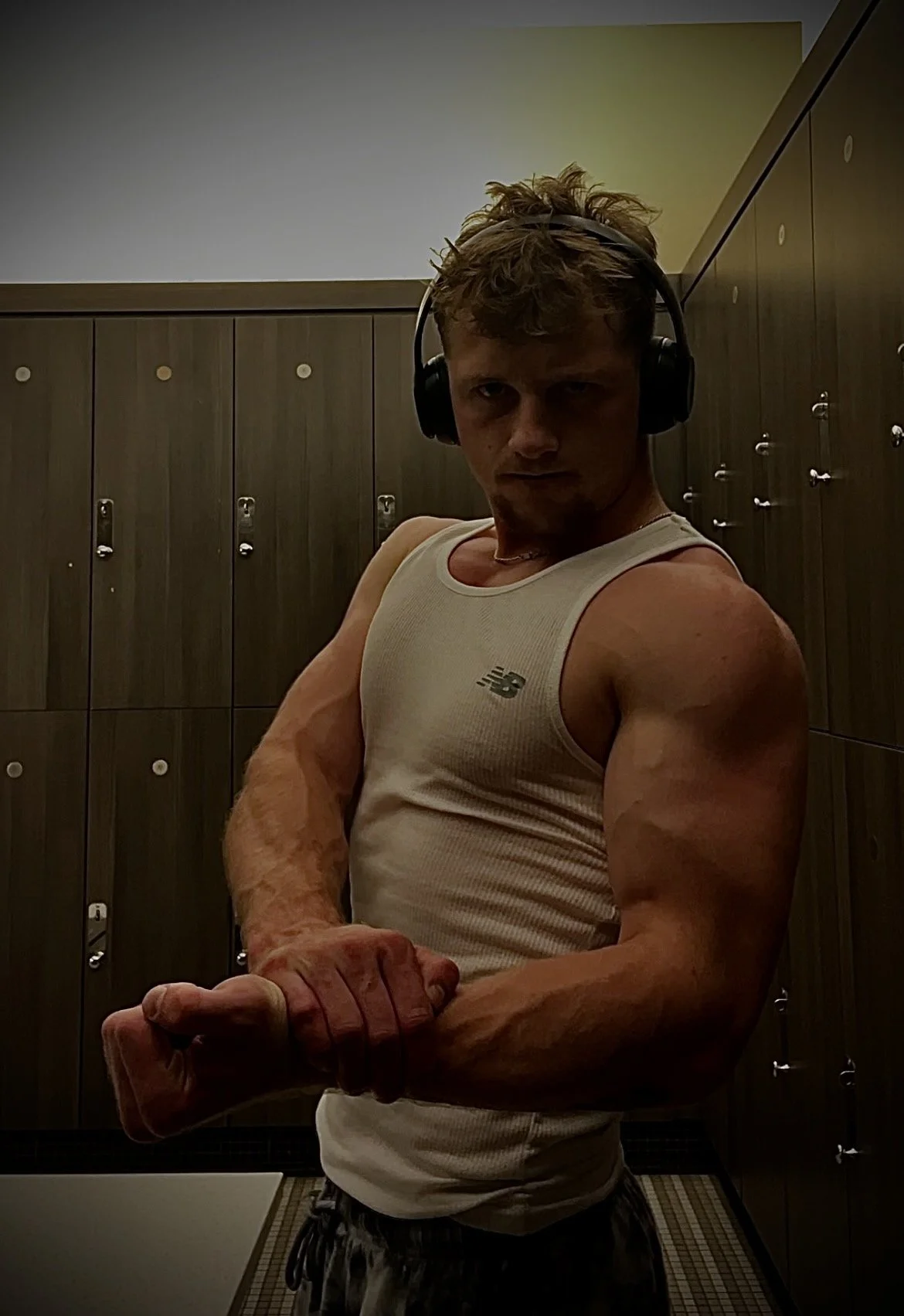 A muscular young man in a gym locker room, wearing headphones, flexing his biceps, and looking at the camera.