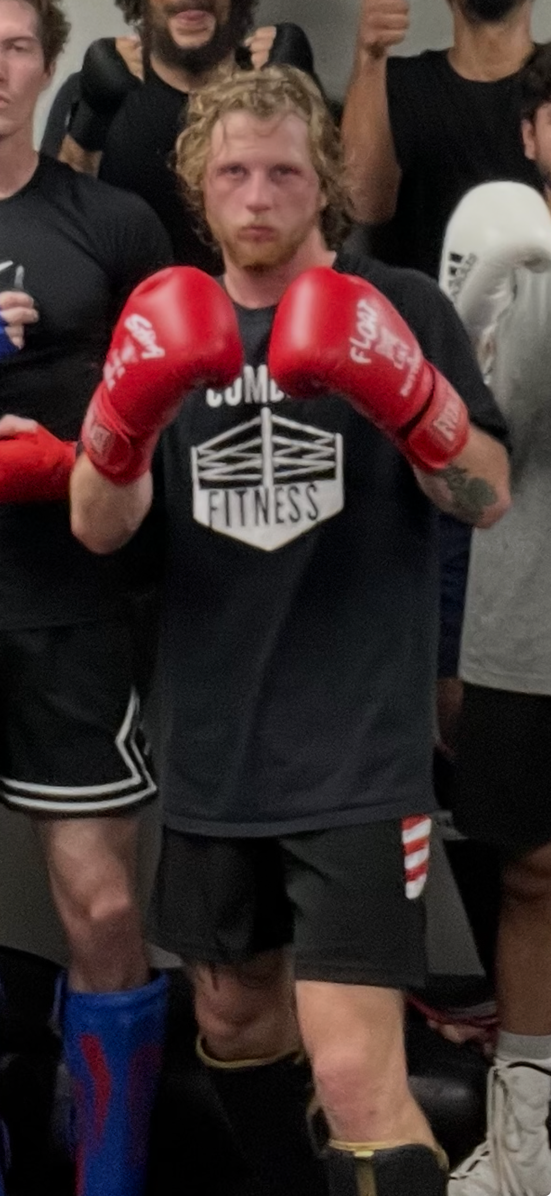 A man with curly hair, a beard, and red boxing gloves is standing in a gym or training facility, surrounded by other people. He is wearing a dark T-shirt with a fitness logo and raises his fists in a fighting stance.