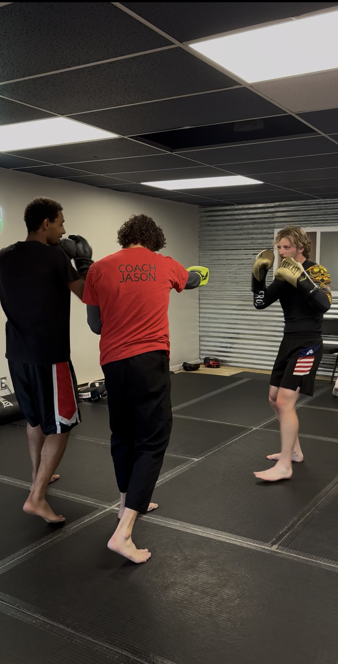 Three men in a boxing gym practicing boxing techniques. One man in black shorts and gloves is facing a man in casual shorts and gloves who is holding focus mitts. The third man, wearing a red shirt with 'COACH JASON' printed on the back, appears to be instructing or coaching.