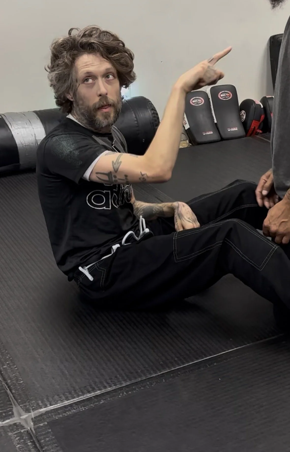 A man with curly hair, tattoos on his arms, and a beard, sitting on the floor of a gym, pointing upward with his right hand. He is wearing a black T-shirt and black pants, with boxing gloves and equipment in the background.