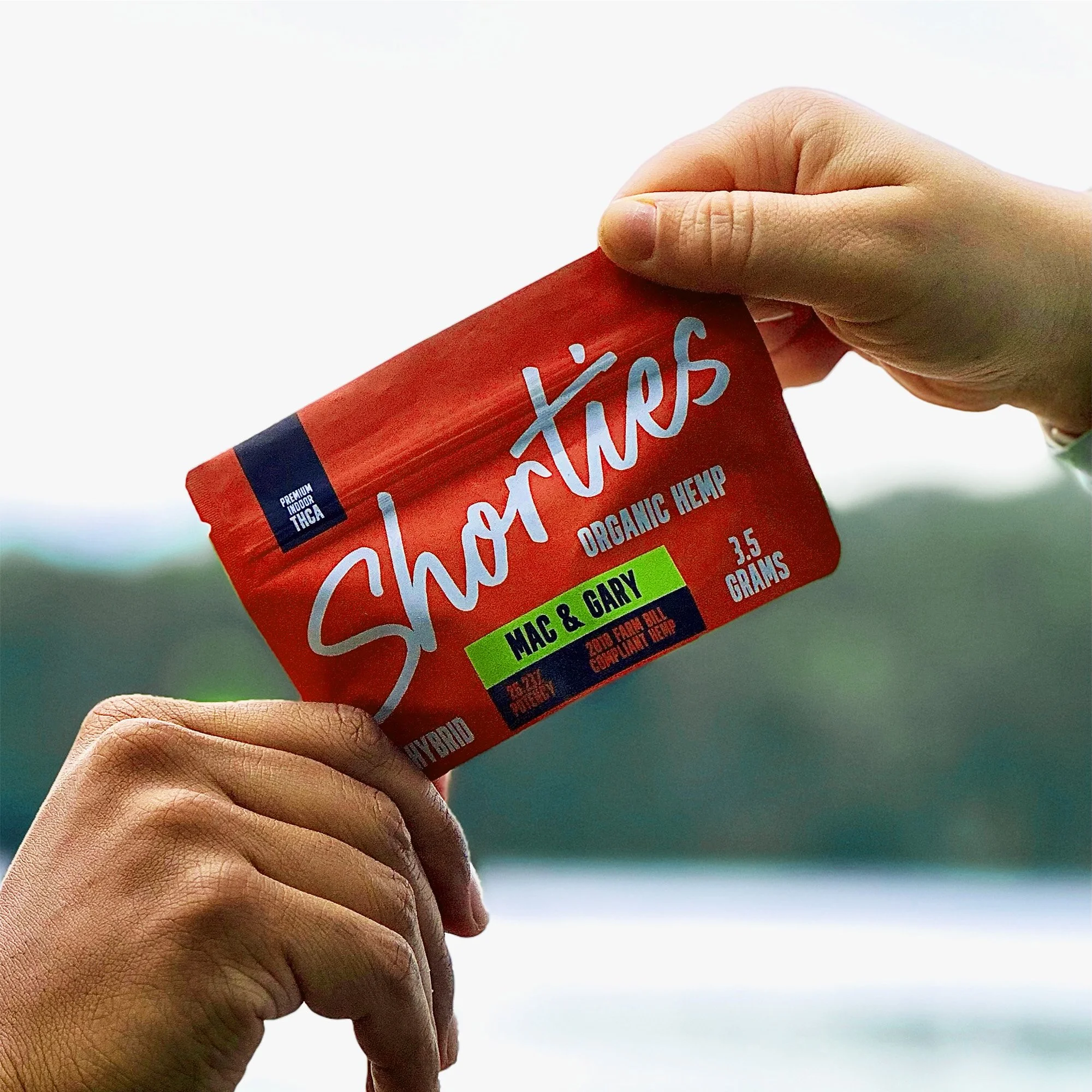 Two hands holding a red package of Shore Ties organic hemp snack with blue and green labels, against a blurry lake or river with trees in the background.