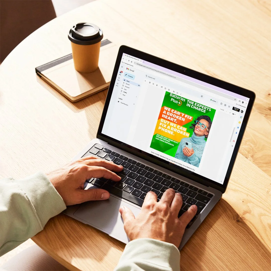 A person working on a laptop at a wooden table, viewing an advertisement for Batteries Plus in an email. There is a notebook and a coffee cup on the table.