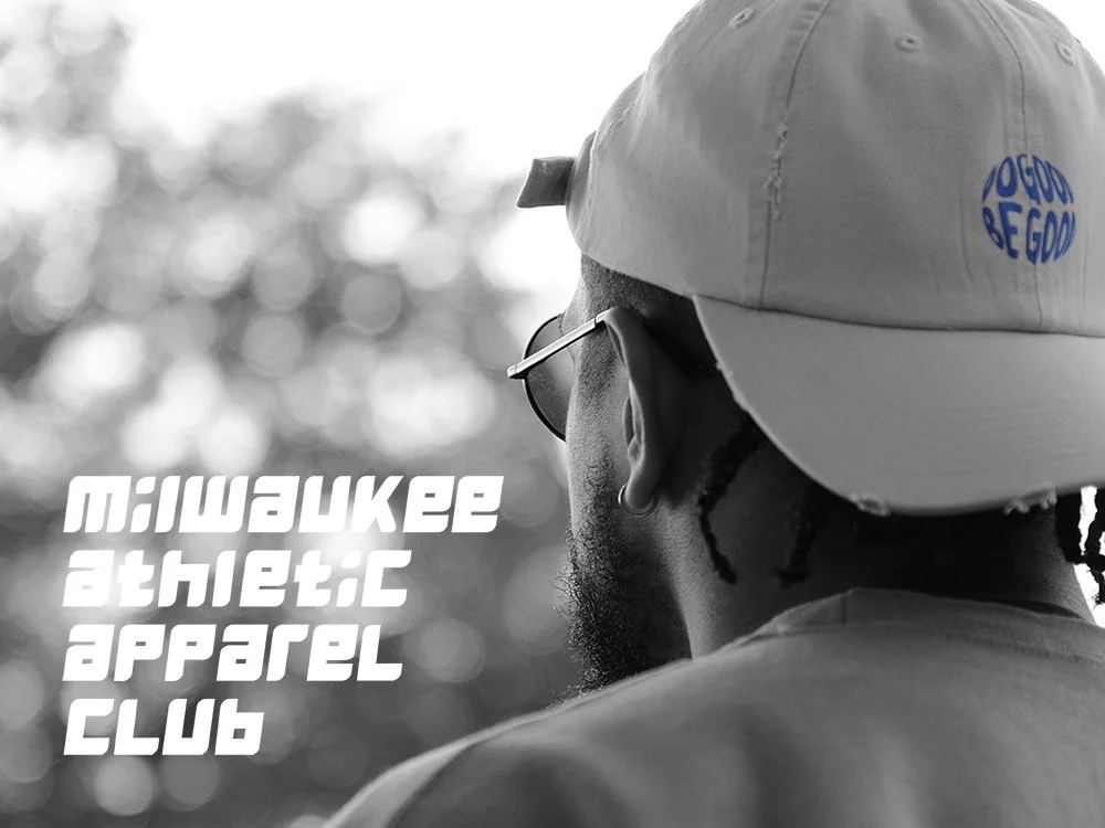 A person wearing a cap with the logo "Good Be Good," sunglasses, and earrings, seen from behind, with the text "Milwaukee Athletic Apparel Club" on the left side of the image, in front of a blurred outdoor background.