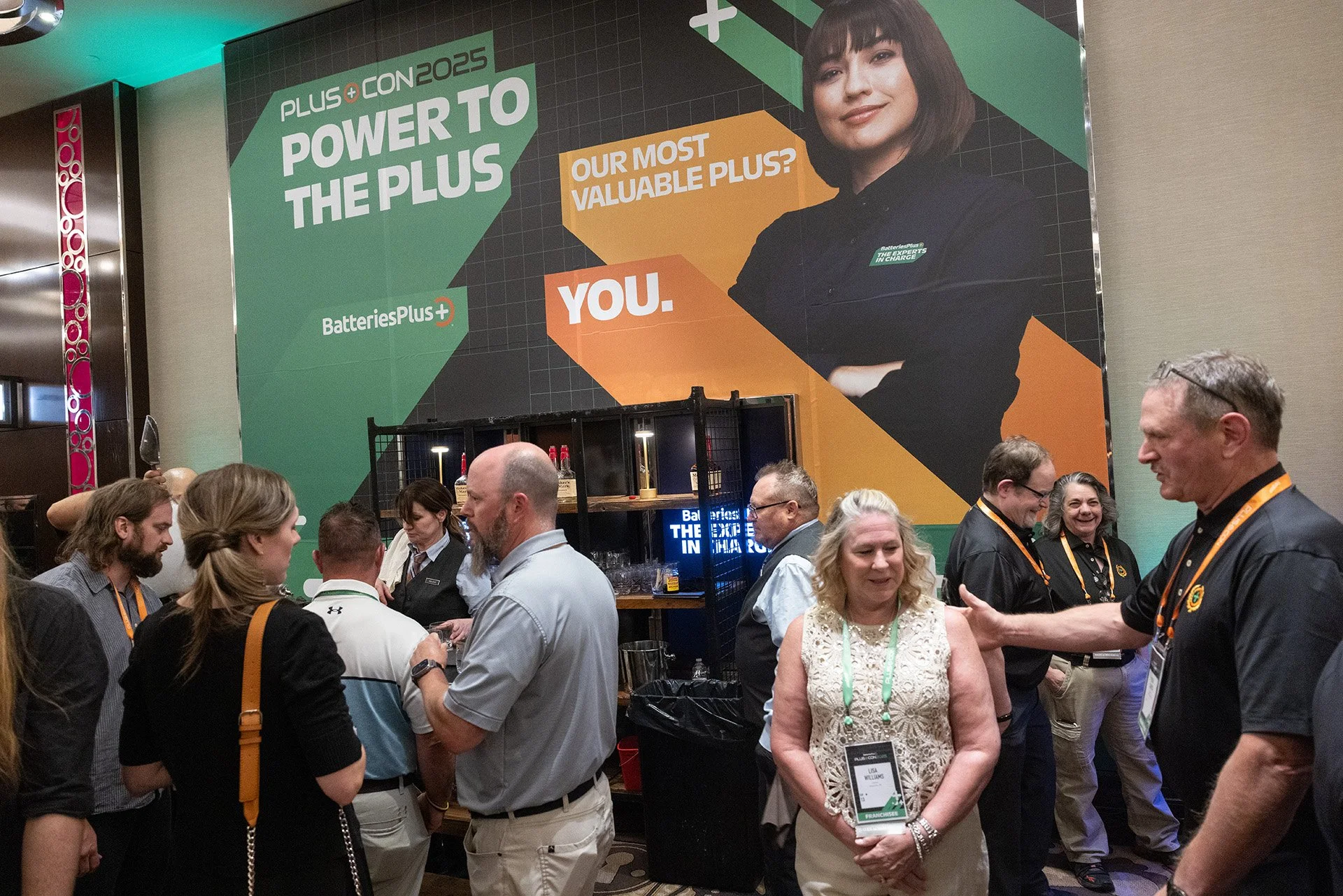 Group of people networking at a conference, with large banners in the background displaying the text "PLUS+ CON 2025 POWER TO THE PLUS" and "BatteriesPlus+".