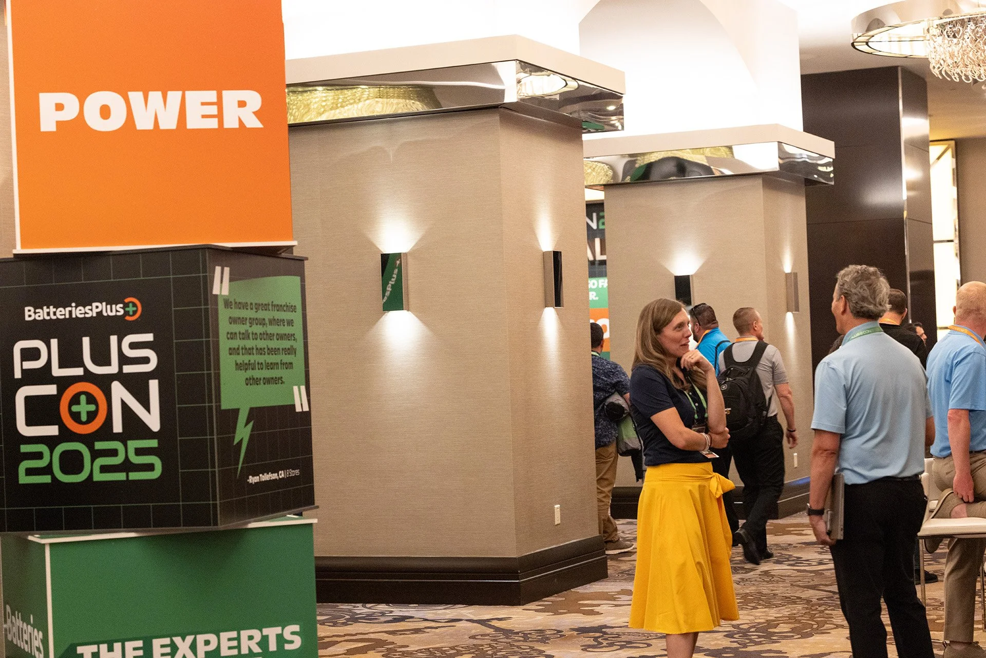 People attending the BatteriesPlus CON 2025 conference in a hotel lobby. A woman in a yellow skirt and navy top is speaking with a man holding a laptop. Conference signs and decorative lighting are visible.