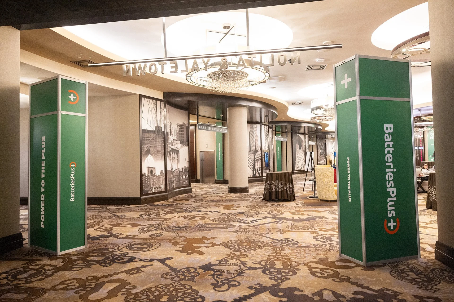 Interior view of a conference hall or hotel lobby with green signage for BatteriesPlus and power outlets, with patterned carpet, decorative wall murals, and ceiling chandeliers.