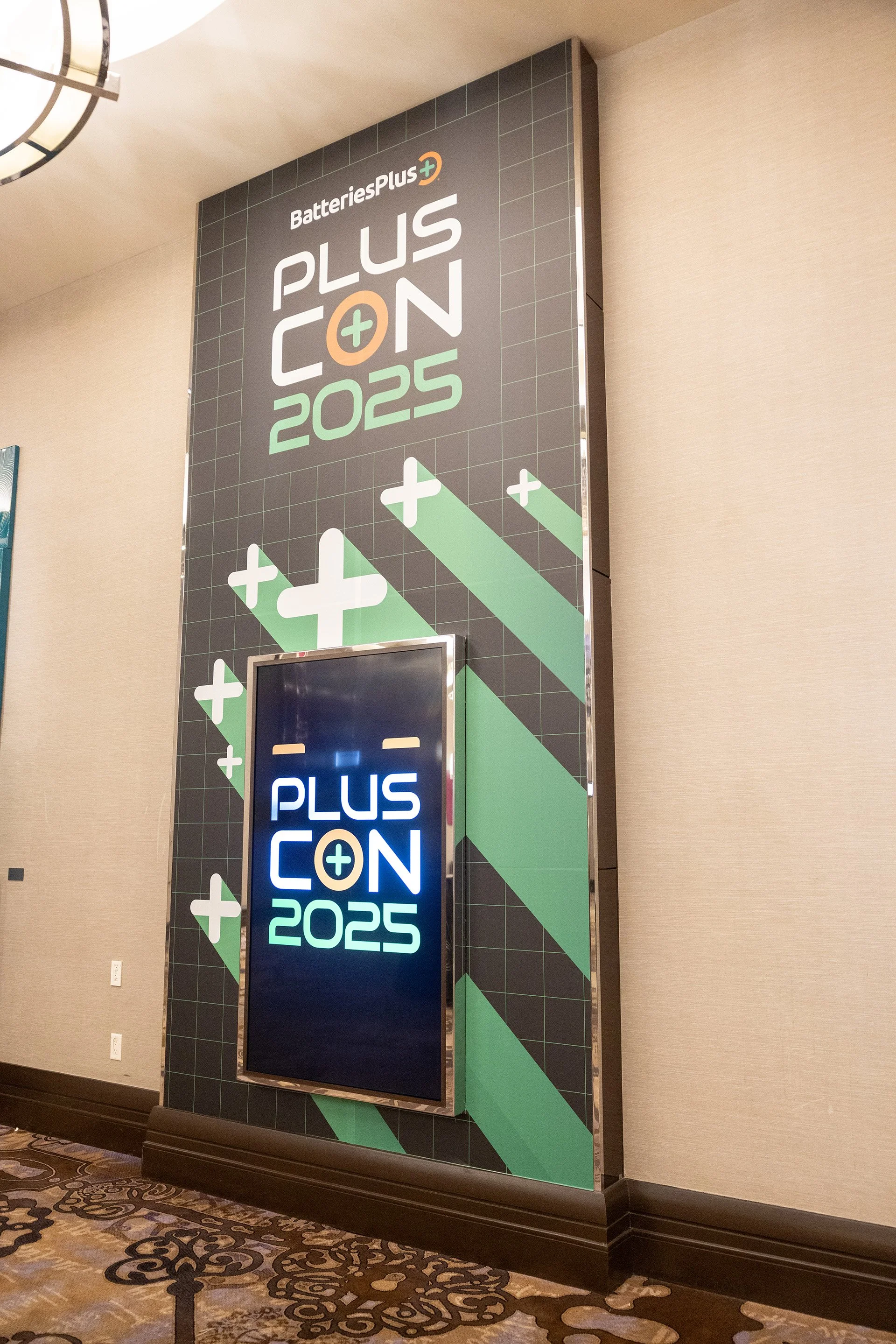 A display wall for BatteriesPlus with the PlusCon 2025 logo, featuring a digital screen showing the same logo in bright neon colors, in an indoor conference or event space.