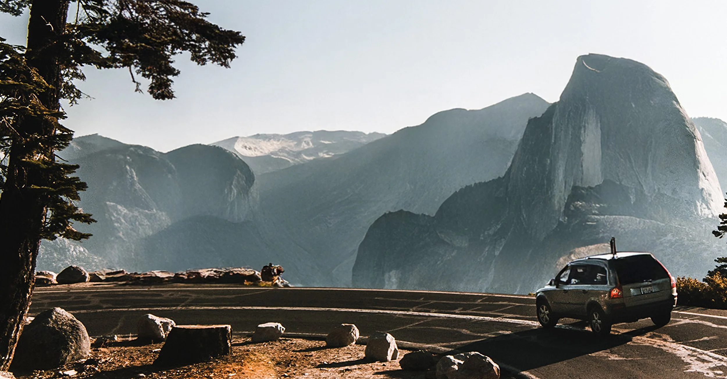 A scenic mountain view with a large rock formation, a parked car, trees in the foreground, and misty mountains in the background.