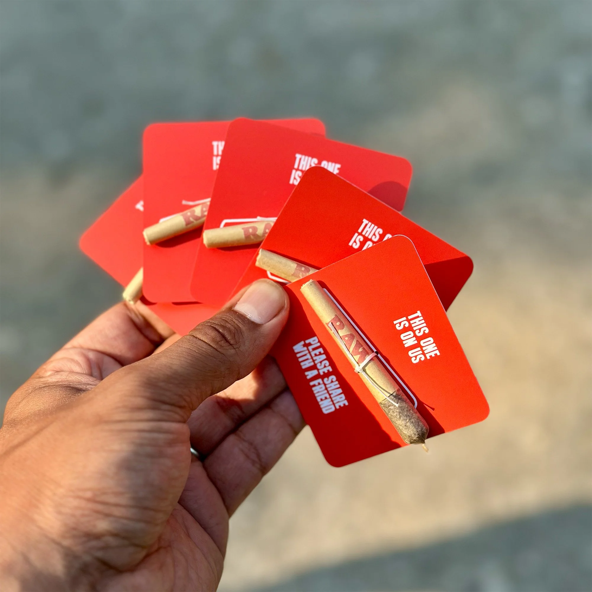 A hand holding four red cardboard holders each containing rolled cigarettes or joints, with a blurred background that appears to be a sandy or beach environment.
