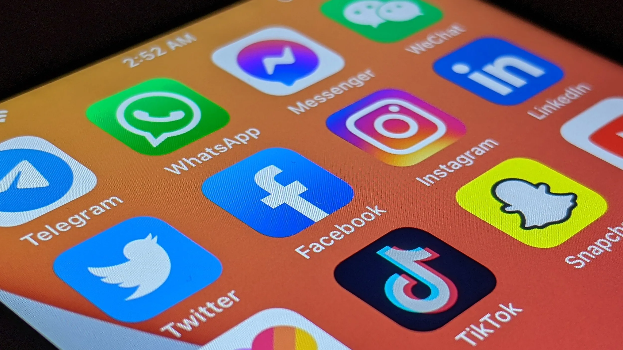 Close-up of a smartphone screen displaying various social media app icons on an orange background, including WhatsApp, Messenger, LinkedIn, Instagram, Facebook, Twitter, TikTok, and Snapchat.