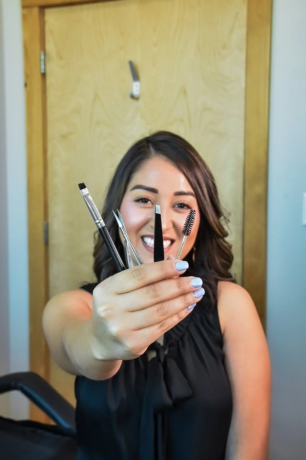 A woman smiling and holding a handful of makeup tools, including brushes, tweezers, and a spoolie, in front of a wooden door.